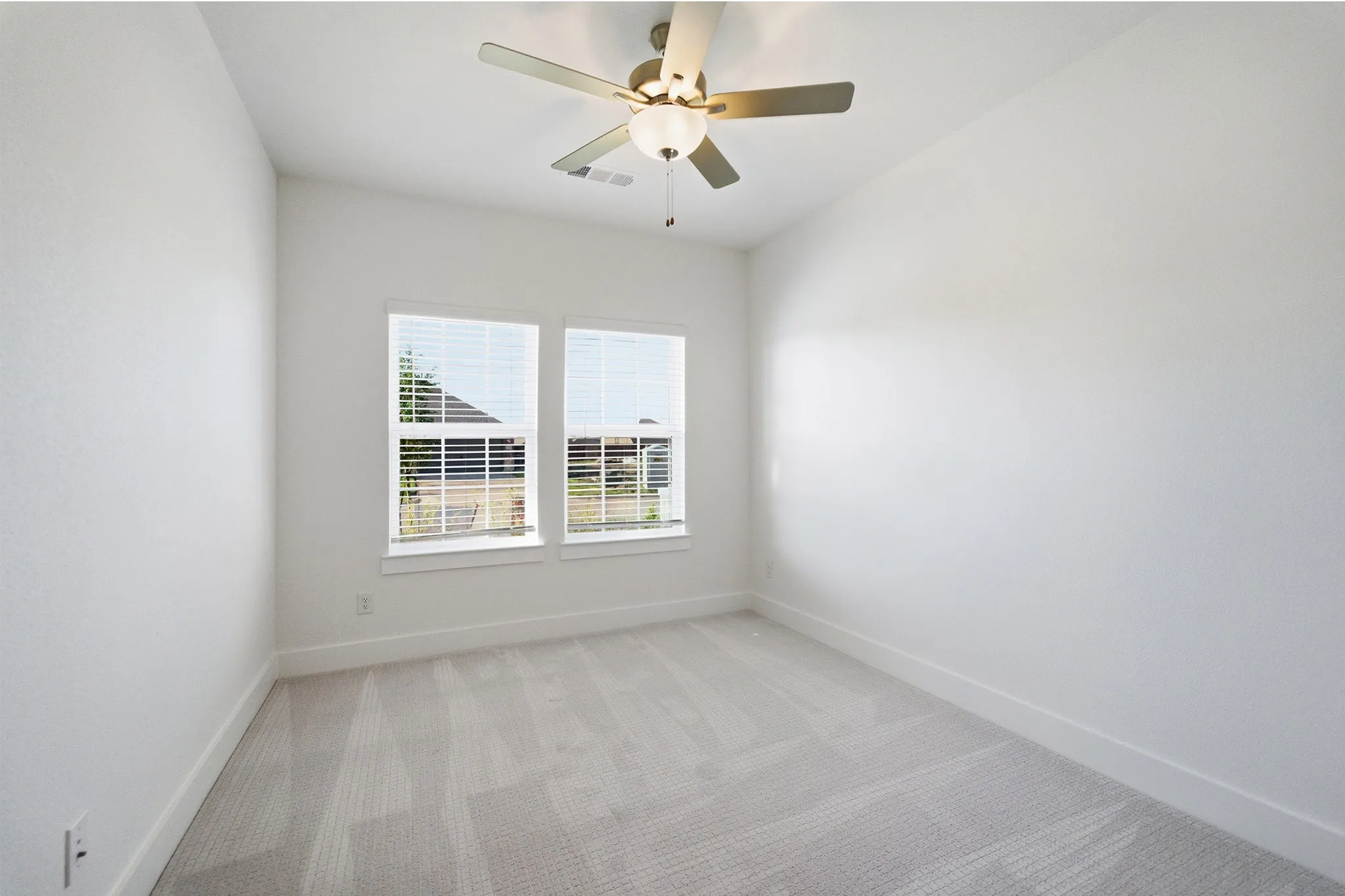 Bright room featuring two windows with blinds, light gray patterned carpet, white walls, and a ceiling fan with integrated lighting