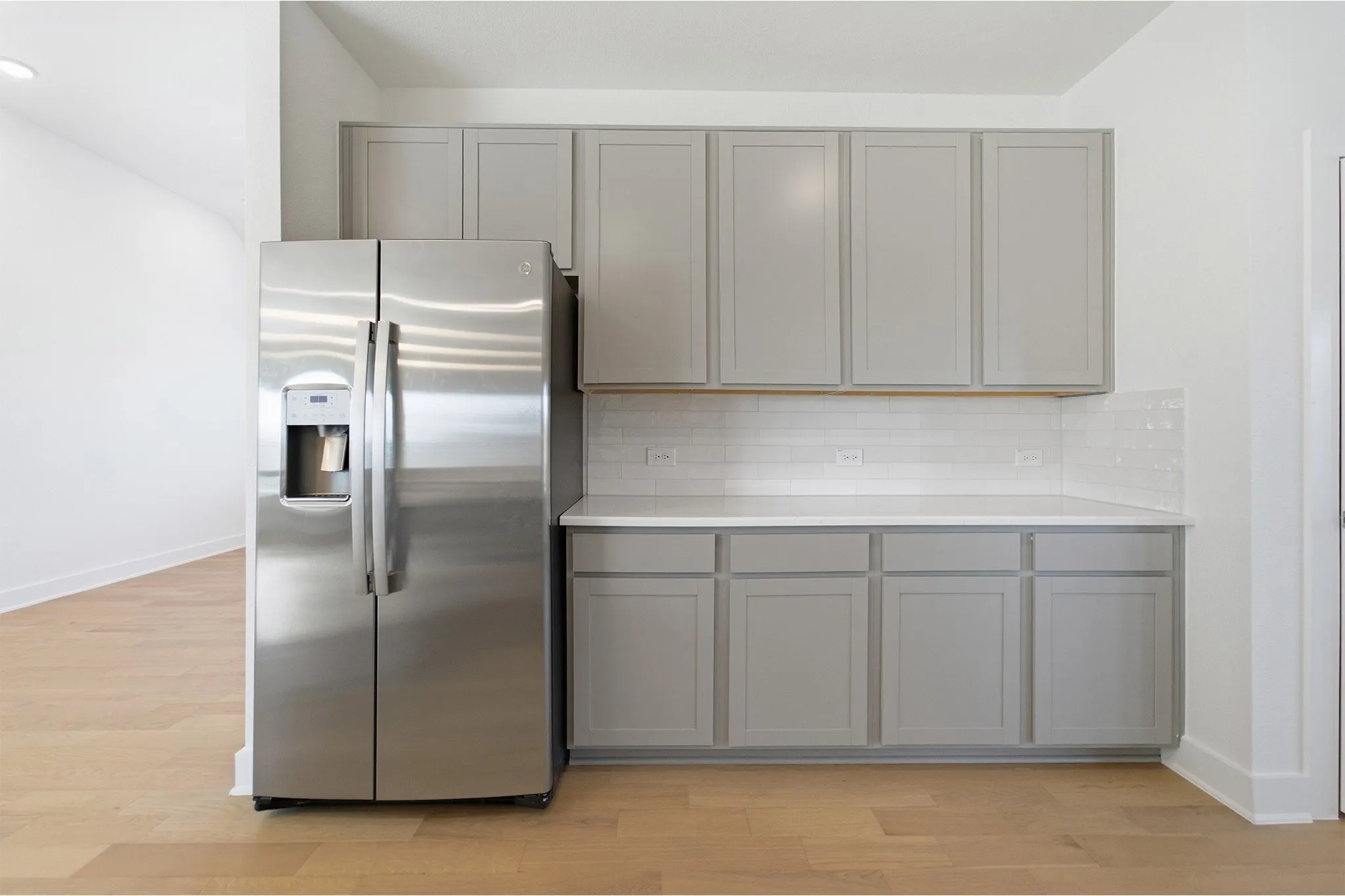 Stainless steel side-by-side refrigerator, light gray shaker cabinetry, white subway tile backsplash, white countertops, and wood-finish flooring