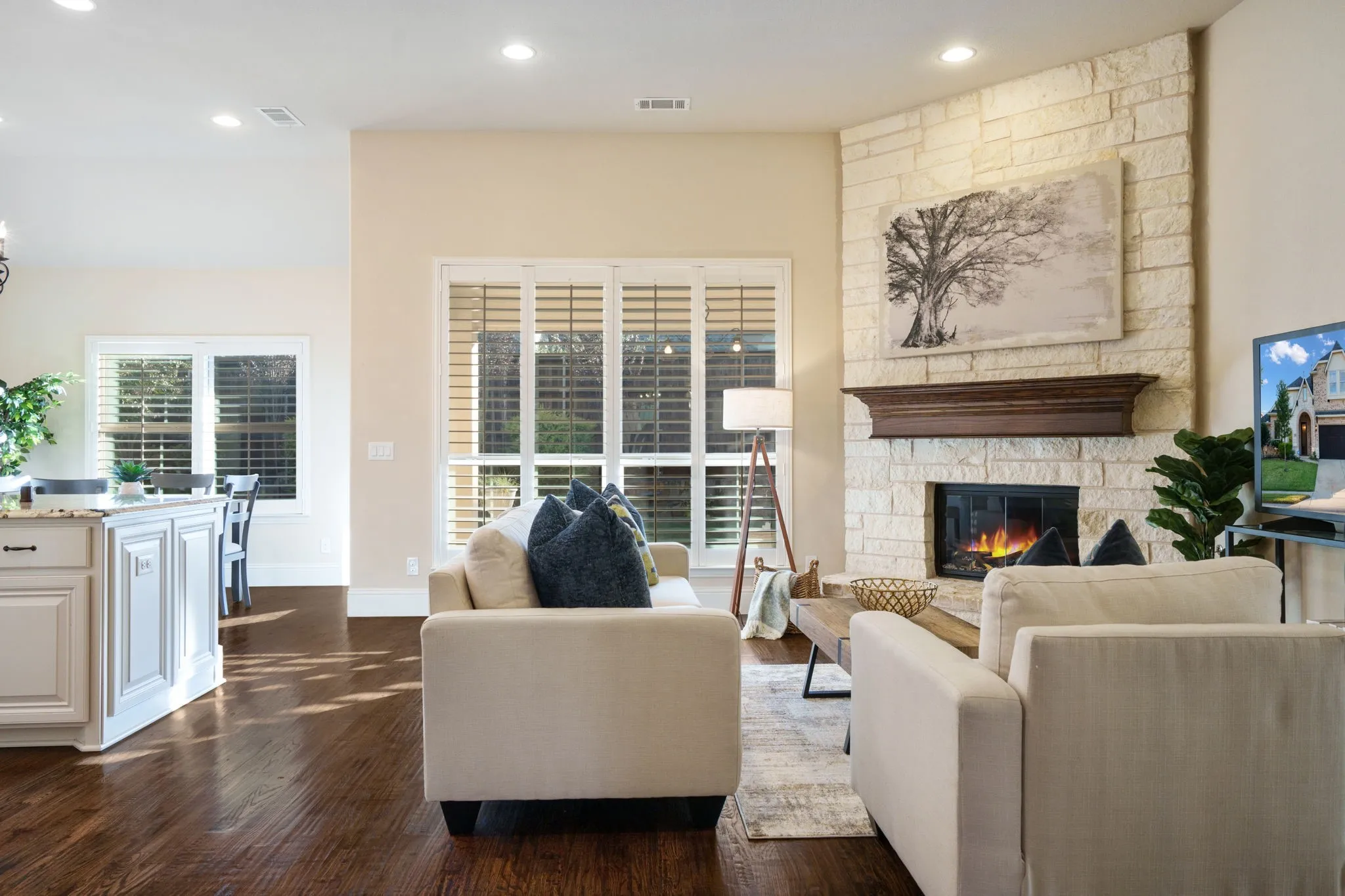 Open-concept living room with stone fireplace, plantation shutters, and views into the kitchen and breakfast area.