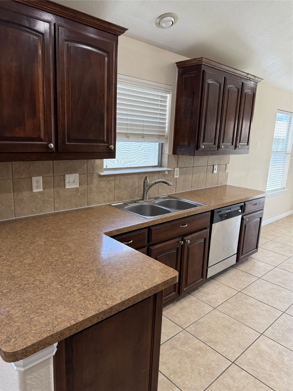 Kitchen with dark brown cabinetry, dishwashing machine, backsplash, light tile patterned floors, and plenty of natural light