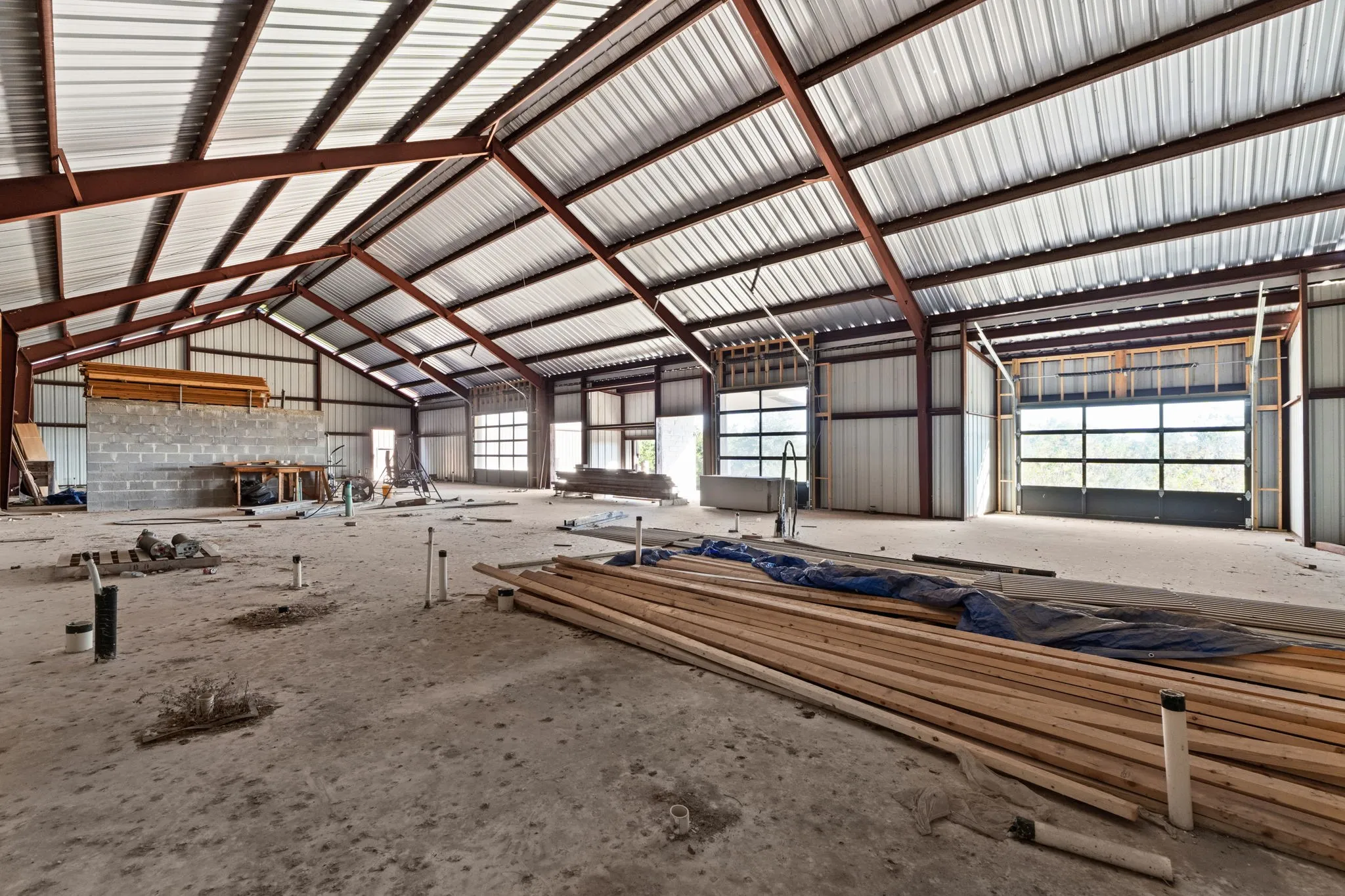 Expansive unfinished interior featuring a vaulted metal ceiling, exposed structural beams, multiple glass-paneled roll-up doors, and visible plumbing rough-ins