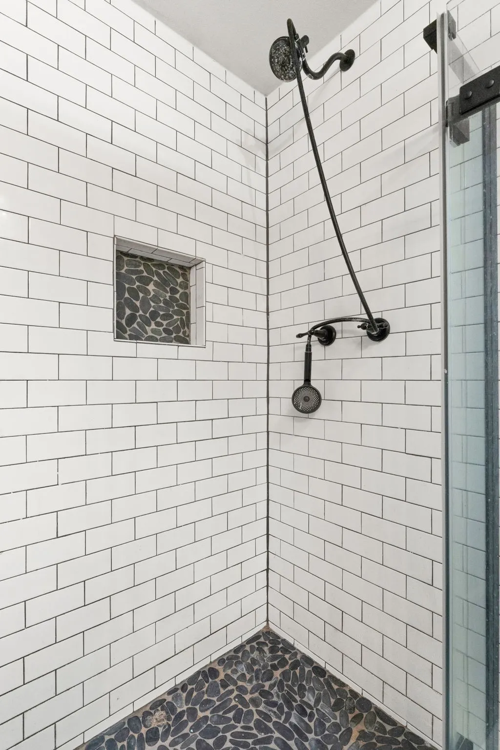 Corner shower featuring white subway tile walls, a pebble stone floor, and a built-in shower niche with pebble stone backing
