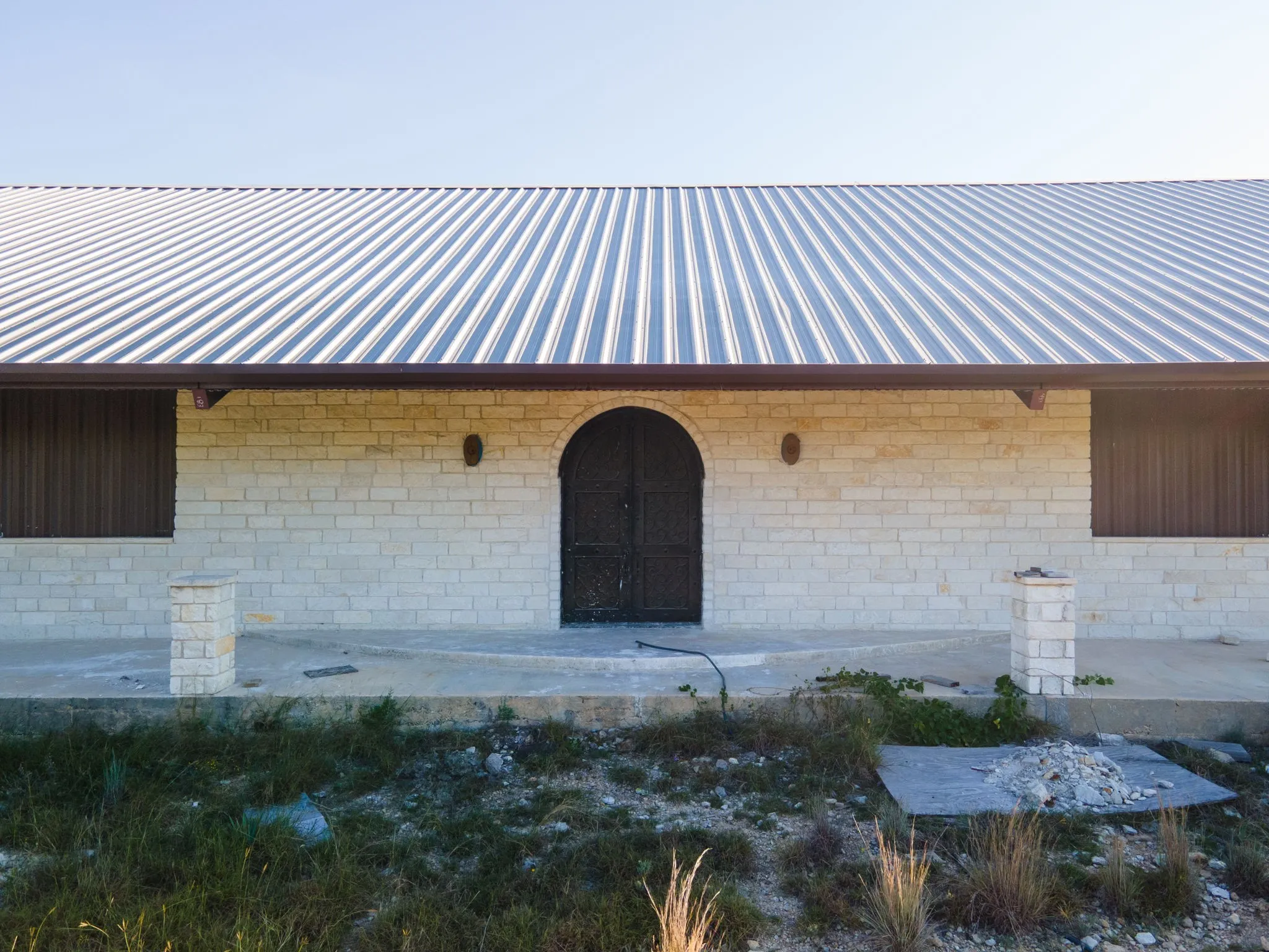 Light-colored brick facade featuring an arched entry, a corrugated metal roof, and a concrete foundational slab