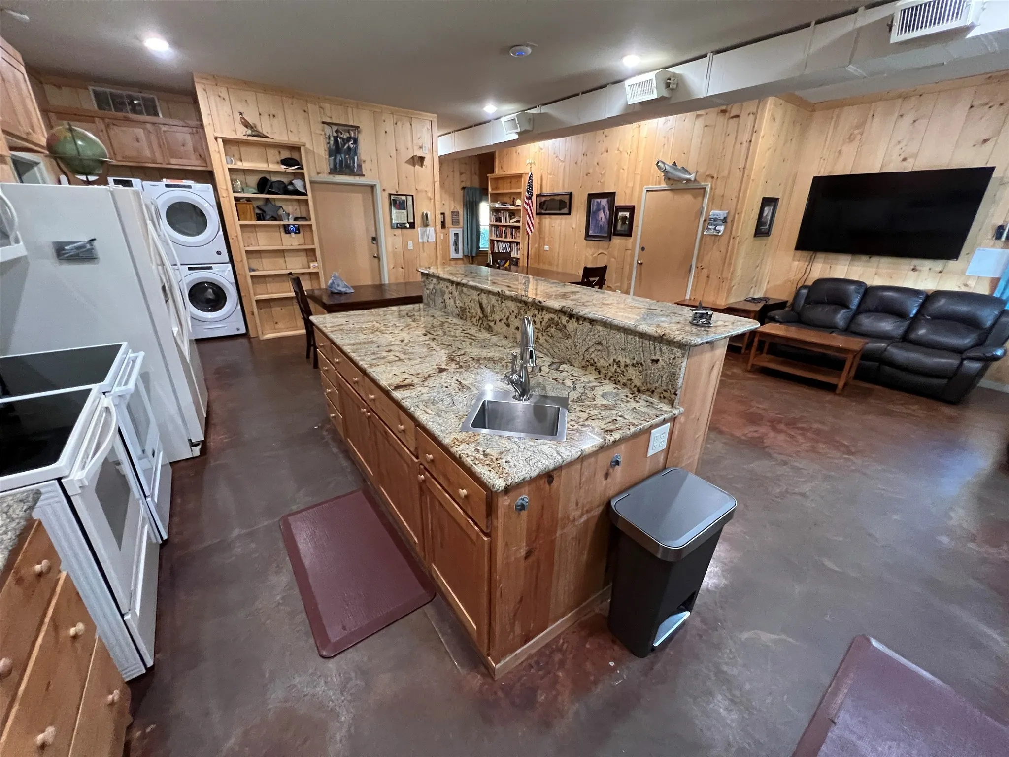 Kitchen featuring concrete flooring, brown cabinets, light stone counters, white appliances, and stacked washing machine and dryer
