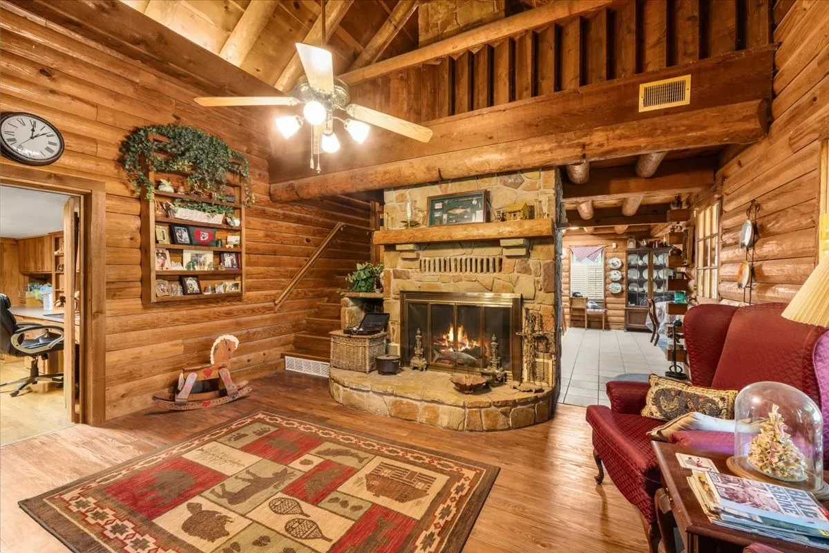 Living area featuring high vaulted ceiling, log walls, a stone fireplace, wood finished floors, and a ceiling fan, virtual fire