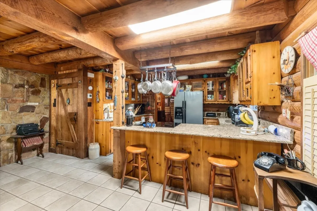 Kitchen with a kitchen bar, light tile patterned floors, stainless steel refrigerator with ice dispenser, glass insert cabinets, and a wooden ceiling with exposed beams