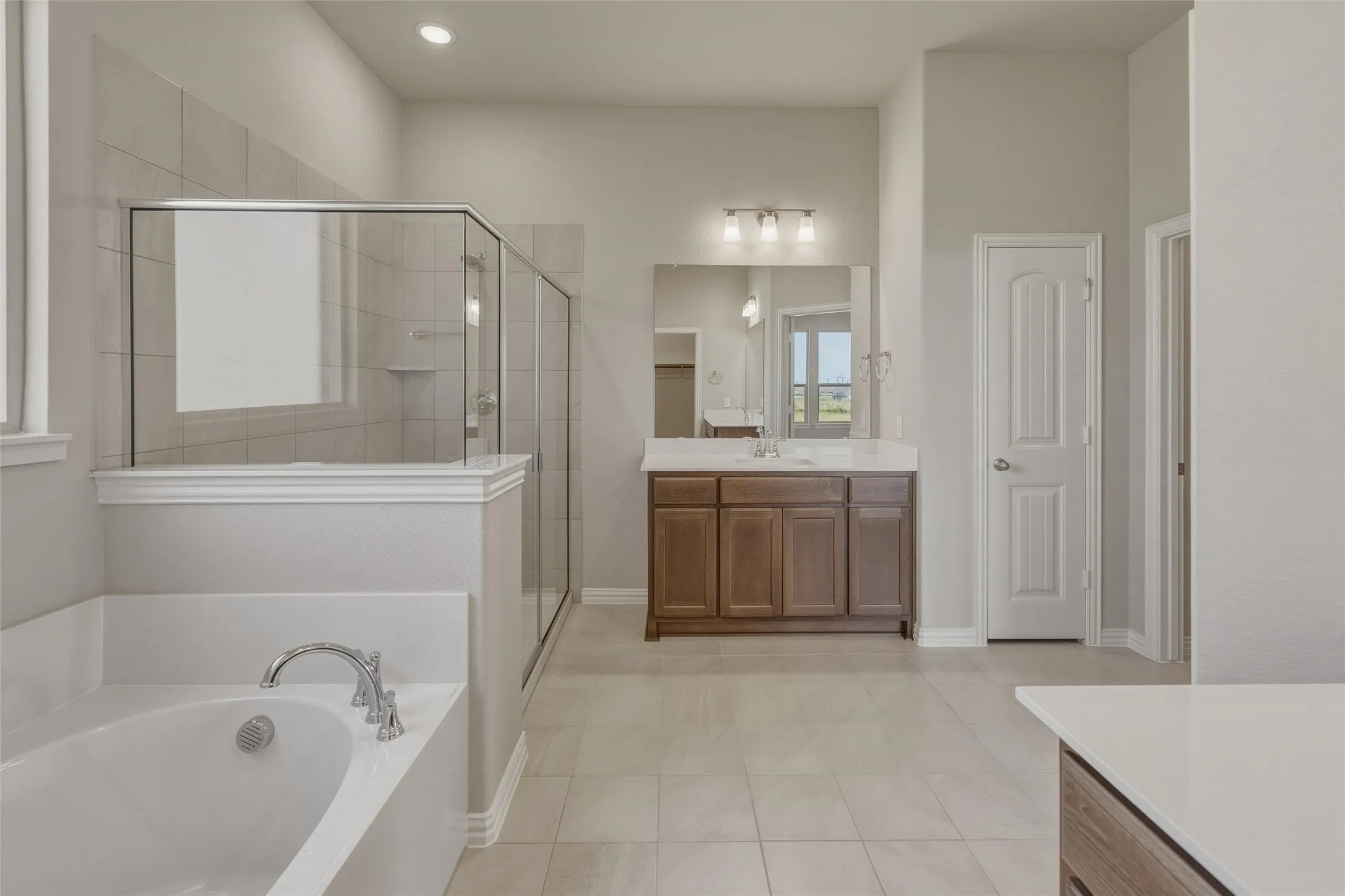 Spacious bathroom featuring a built-in soaking tub with chrome fixtures, a separate glass-enclosed shower with tiled walls, and a wood-finish vanity with a white countertop and rectangular mirror