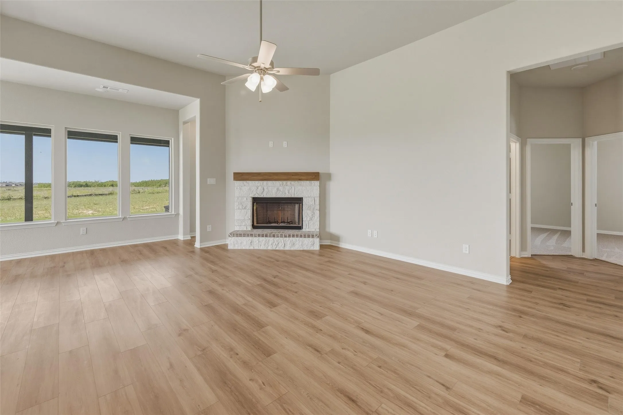 Spacious living area featuring wood-finish flooring, a white stone-faced fireplace with wood mantel, a ceiling fan, and multiple large windows overlooking the surrounding landscape