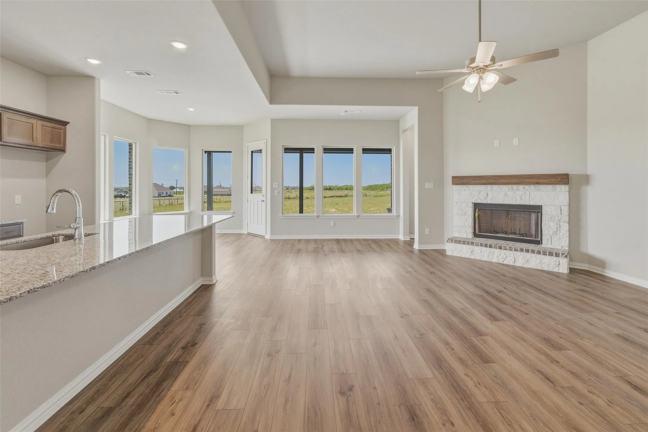 Open-concept living area featuring wood-finish flooring, a white brick fireplace with a wood mantel, a ceiling fan, recessed lighting, and expansive windows