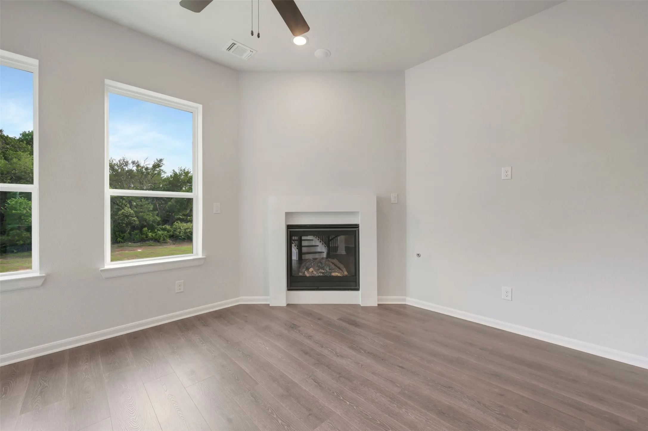 Bright living area featuring a fireplace with a white surround, wood-finish flooring, two large windows, and a ceiling fan