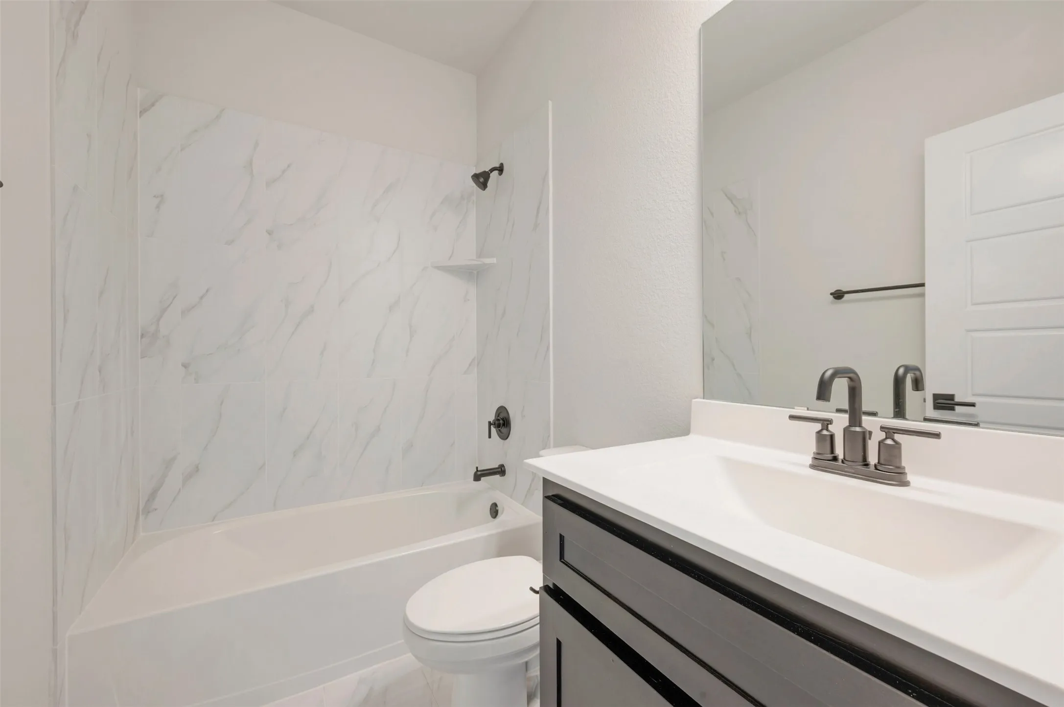 Bathroom featuring a white tub with a white-toned, patterned surround, a dark-toned vanity cabinet, a white countertop with an integrated sink, and dark-toned fixtures