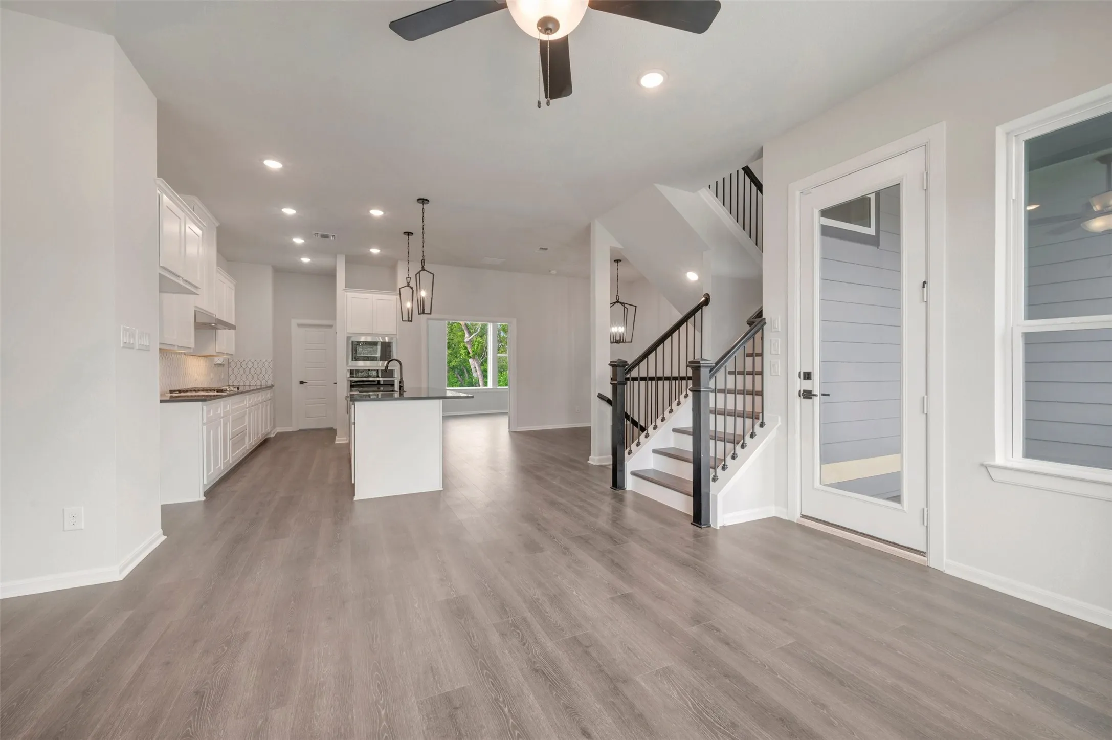 Open-concept living area featuring wood-finish flooring, a contemporary kitchen, a staircase with dark wood newel posts and railings, recessed lighting, and a ceiling fan with integrated light