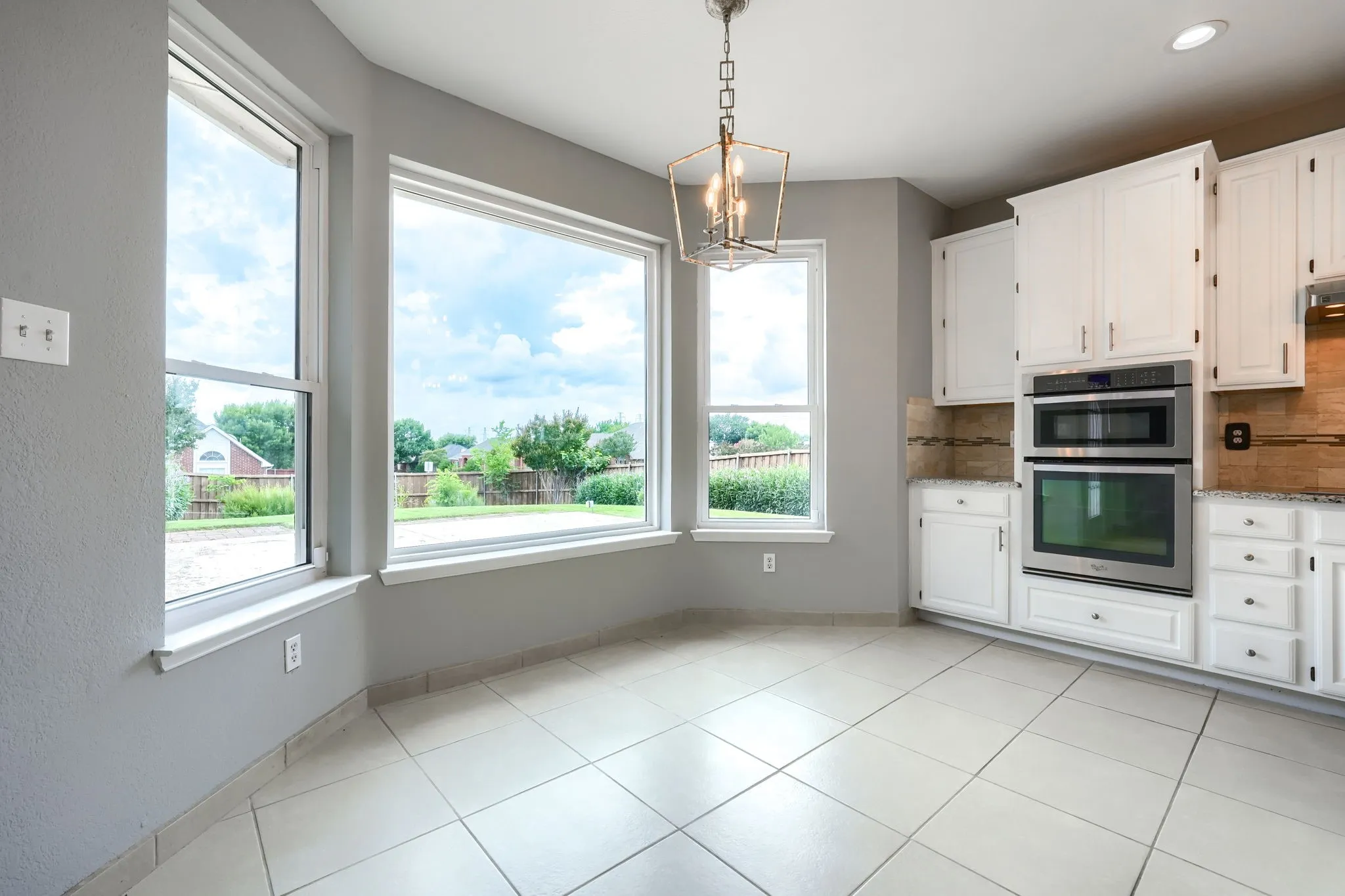 Kitchen dining area featuring white cabinetry, light-toned tiled flooring, stainless steel double ovens, granite countertops, and a decorative pendant light fixture