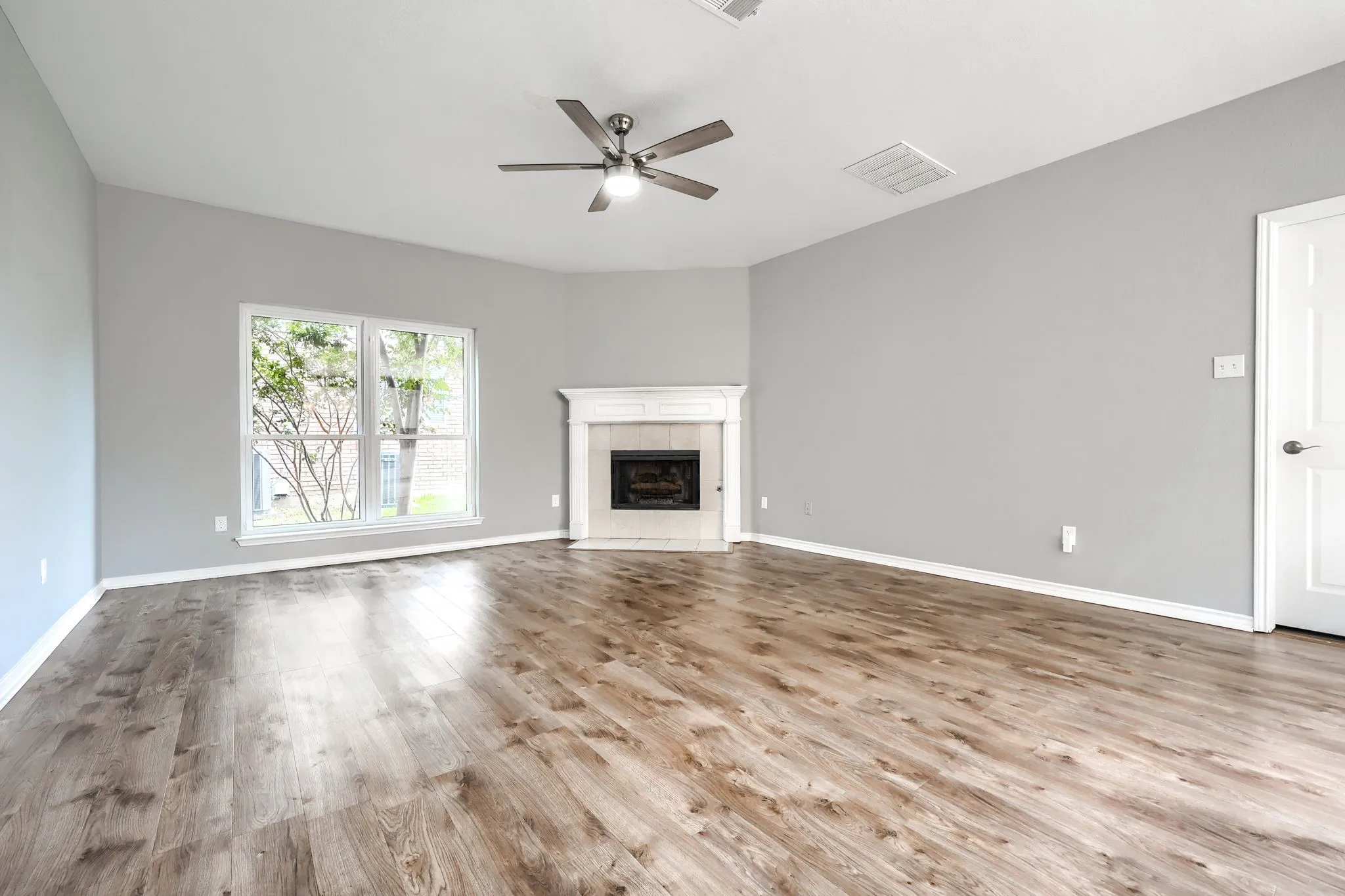 Spacious room featuring wood-finish flooring, a corner fireplace with a white mantel, and a ceiling fan