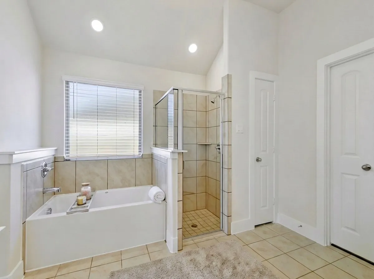 White soaking tub featuring tile surround, frosted window with blinds, and recessed lighting