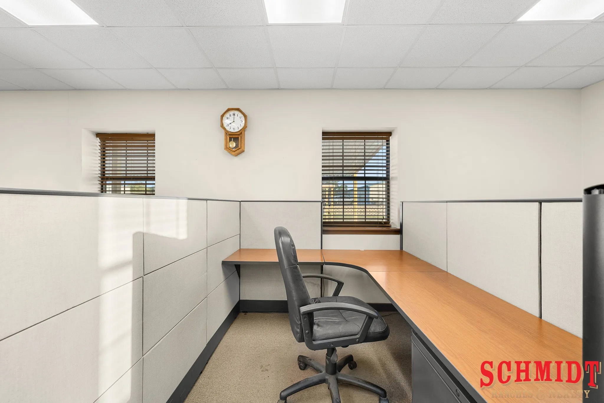 Open-plan office space featuring modular cubicle walls and two windows with wood-finish blinds