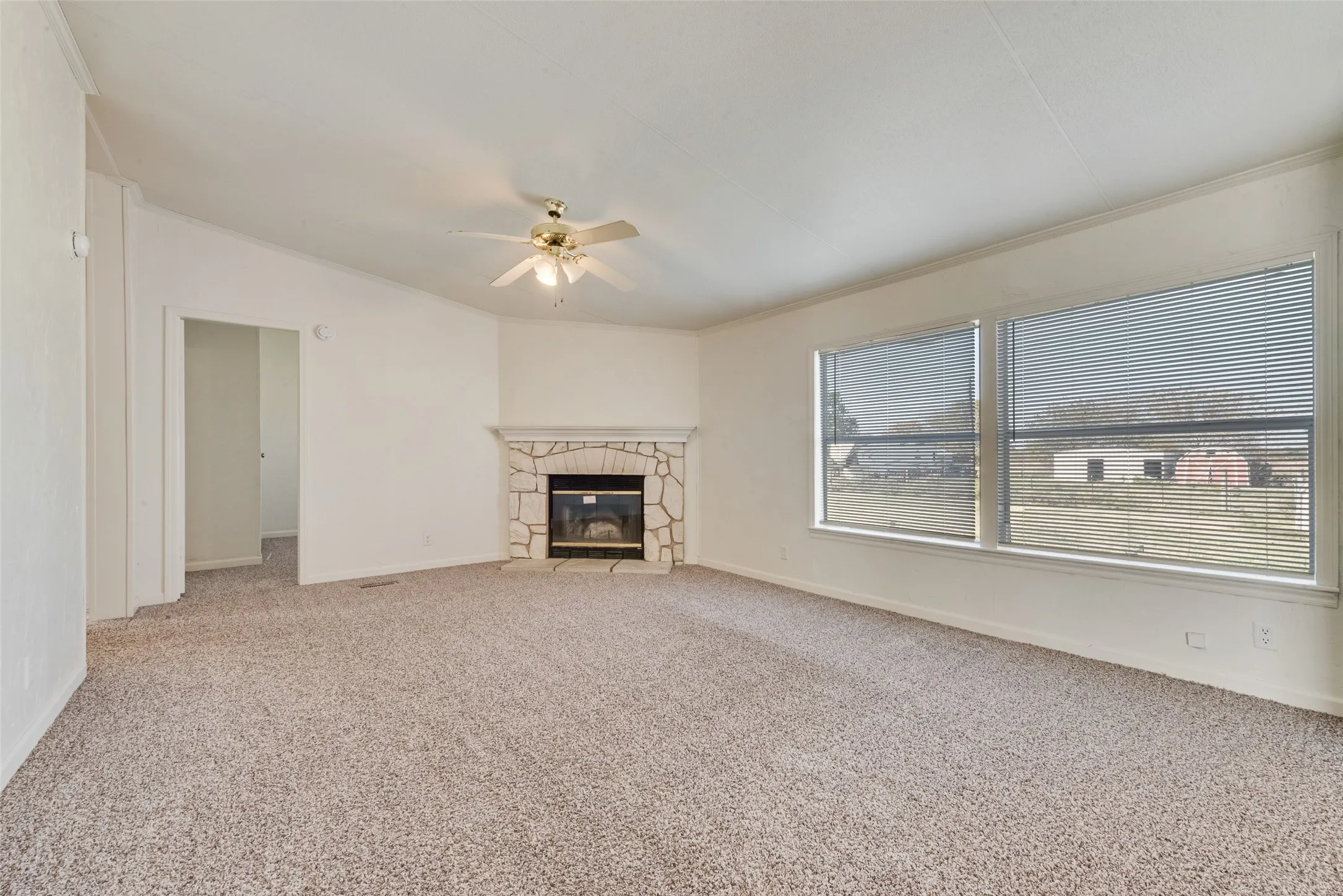Unfurnished living room with light colored carpet, a stone fireplace, crown molding, and ceiling fan