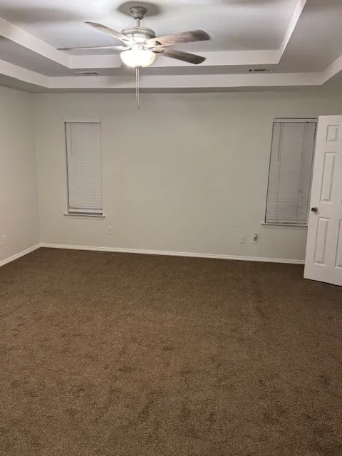Empty room with a raised ceiling, dark carpet, and a ceiling fan