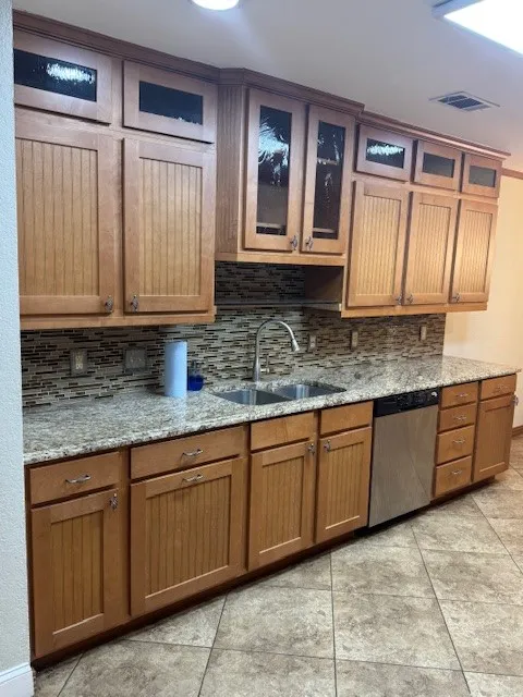 Kitchen featuring light stone countertops, tasteful backsplash, brown cabinetry, dishwasher, and glass insert cabinets