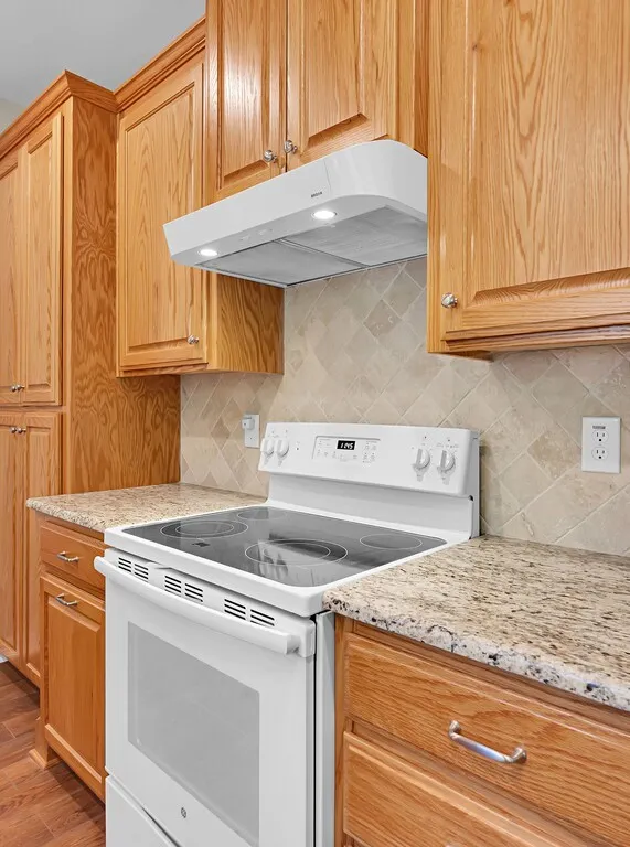 Kitchen with white electric range oven, range hood, backsplash, and light stone counters