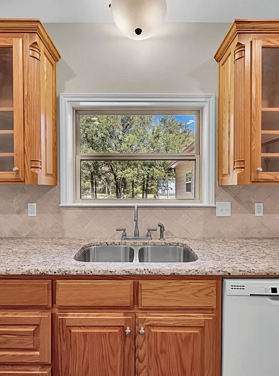 Kitchen featuring backsplash, glass insert cabinets, dishwasher, light stone countertops, and brown cabinets