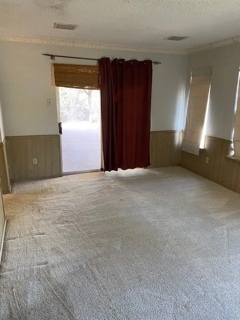 Unfurnished bedroom with a textured ceiling, light colored carpet, wainscoted wall and sliding glass door leading to upper covered deck.