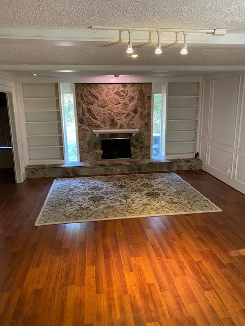 Unfurnished living room featuring a stone fireplace, wood finished floors, track lighting and built in bookshelves.