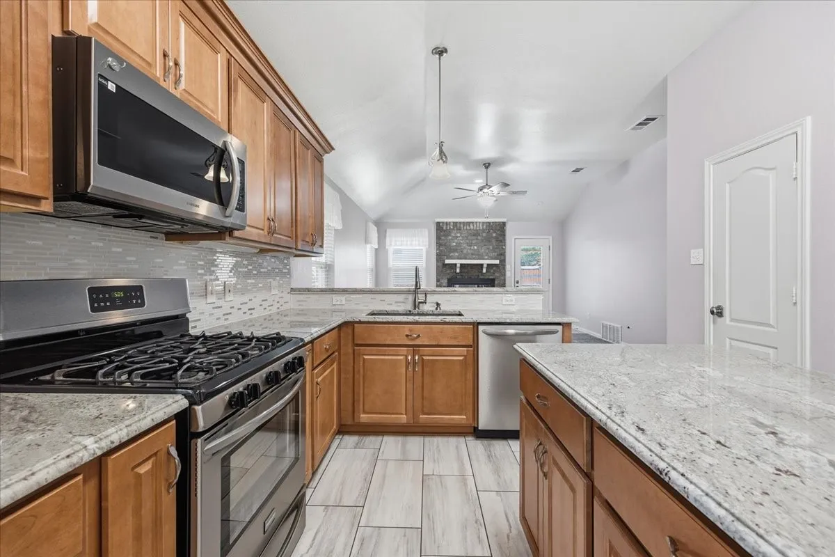 Kitchen featuring brown cabinets, appliances with stainless steel finishes, decorative backsplash, light stone countertops, and lofted ceiling