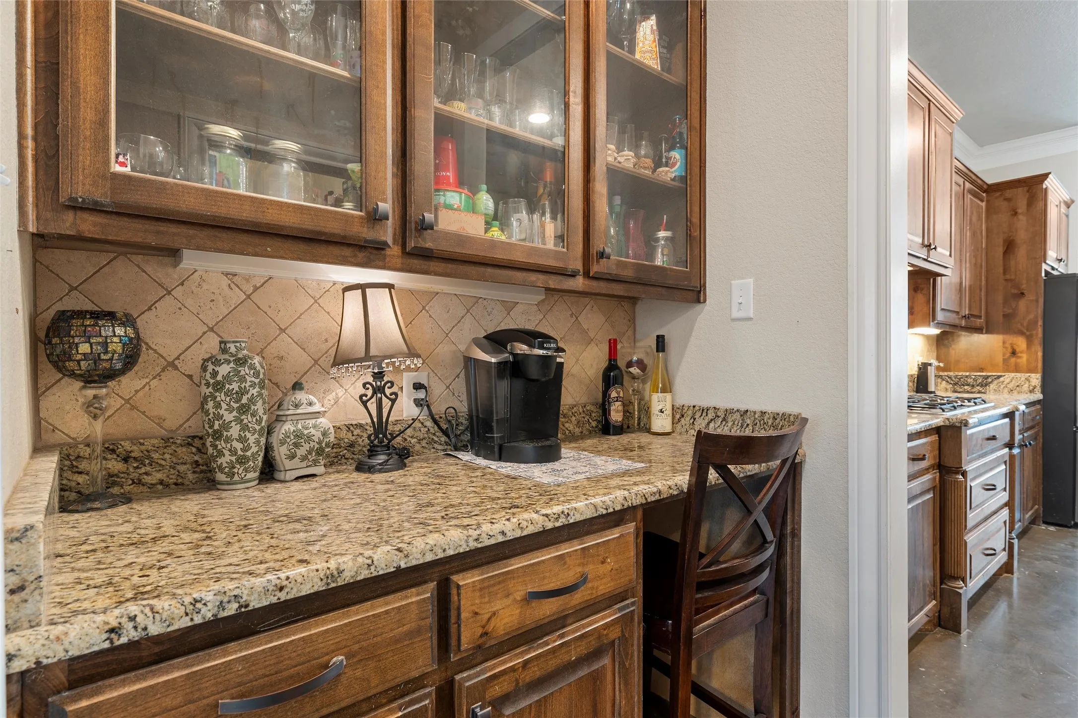 Bar featuring tasteful backsplash, light stone countertops, glass insert cabinets, brown cabinetry, and ornamental molding