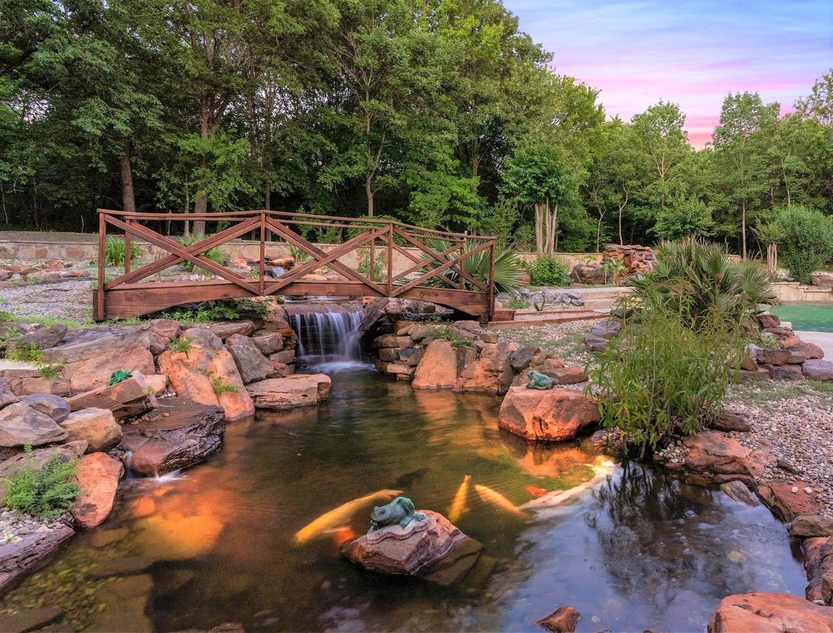 Peaceful Koi Pond with a Tiered Rock Waterfall. A Handcrafted Wooden Bridge Spans Over the Water, Creating a Tranquil Focal Point Surrounded By Lush, Mature Trees and Natural Stone.