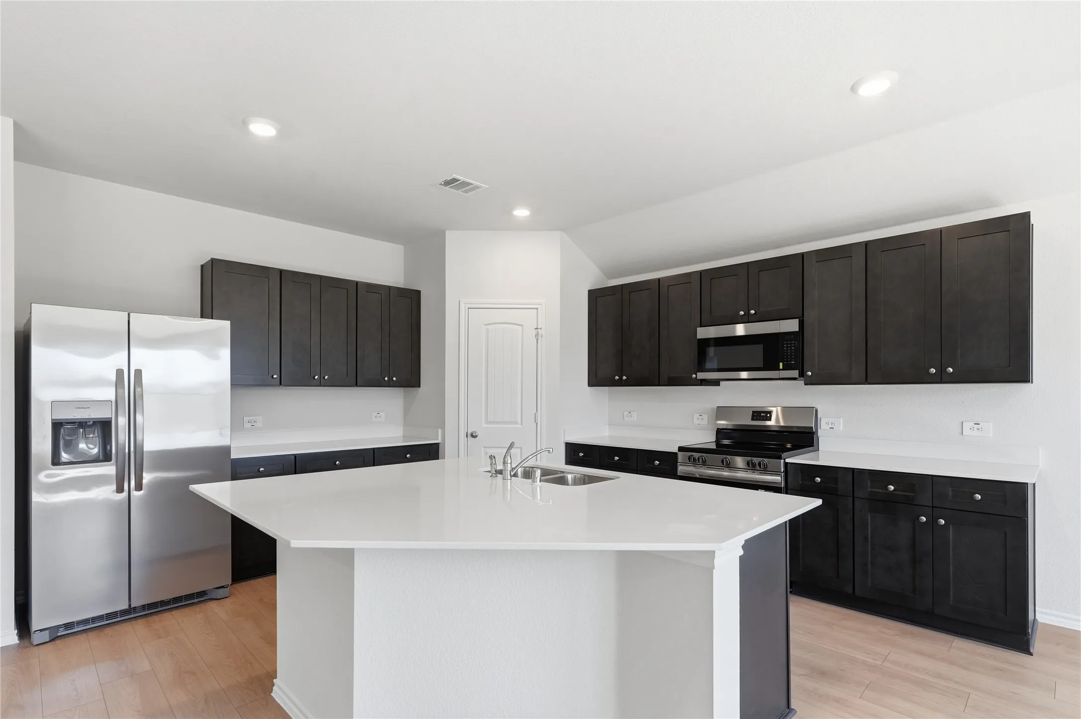 Kitchen featuring a central island with an undermount double-basin sink, stainless steel appliances, dark wood-finish cabinetry, light countertops, and wood-finish flooring