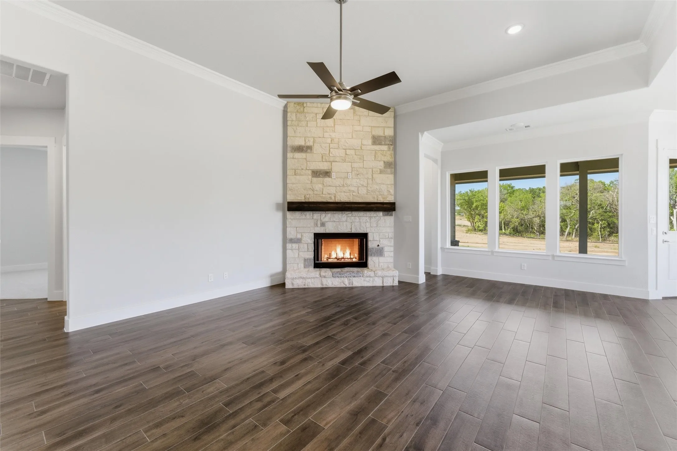 Unfurnished living room with dark wood-style floors, a stone fireplace, a ceiling fan, and ornamental molding