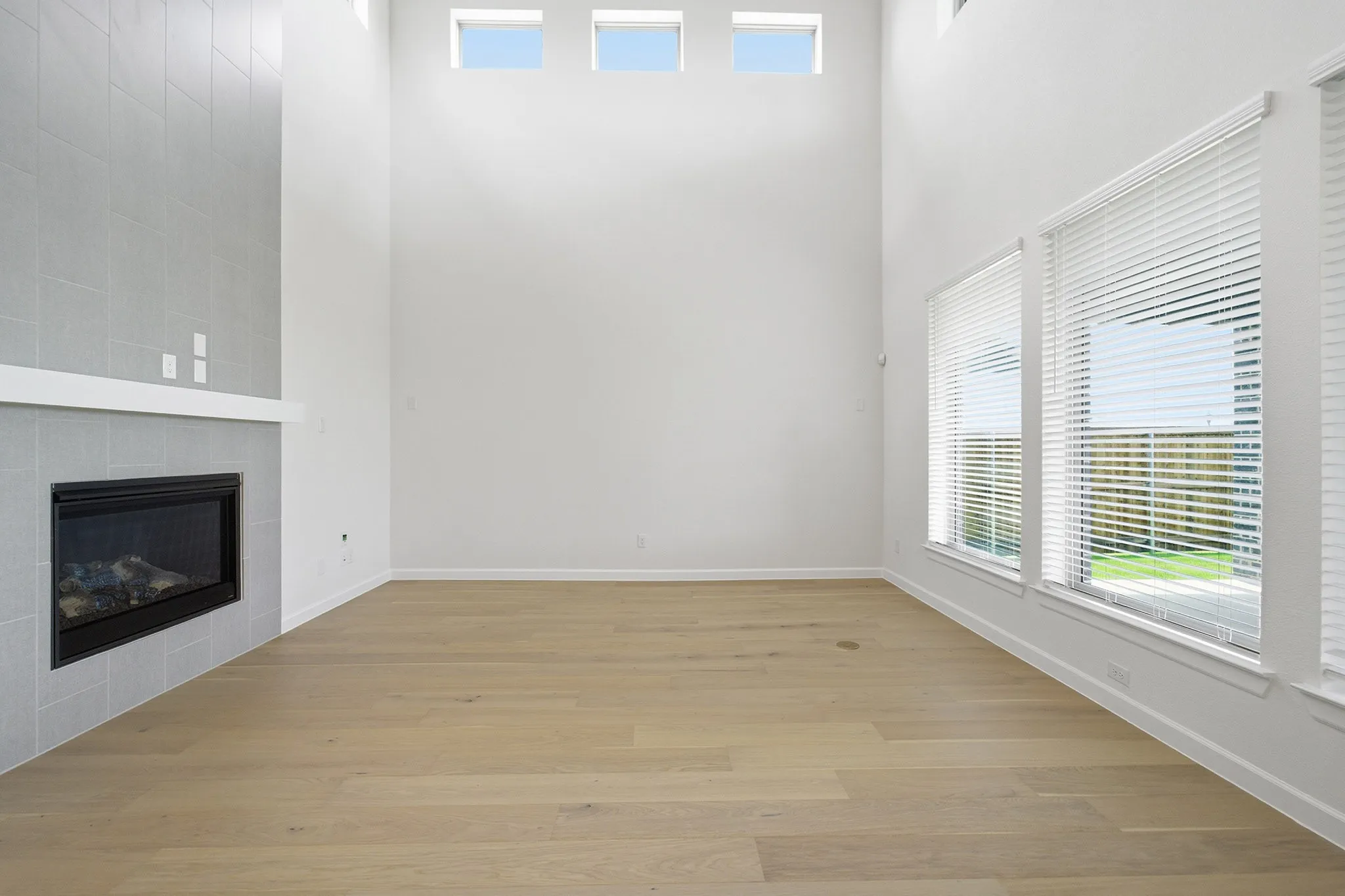 Two-story living area featuring a tiled fireplace with mantel, wood-finish flooring, and multiple windows with blinds