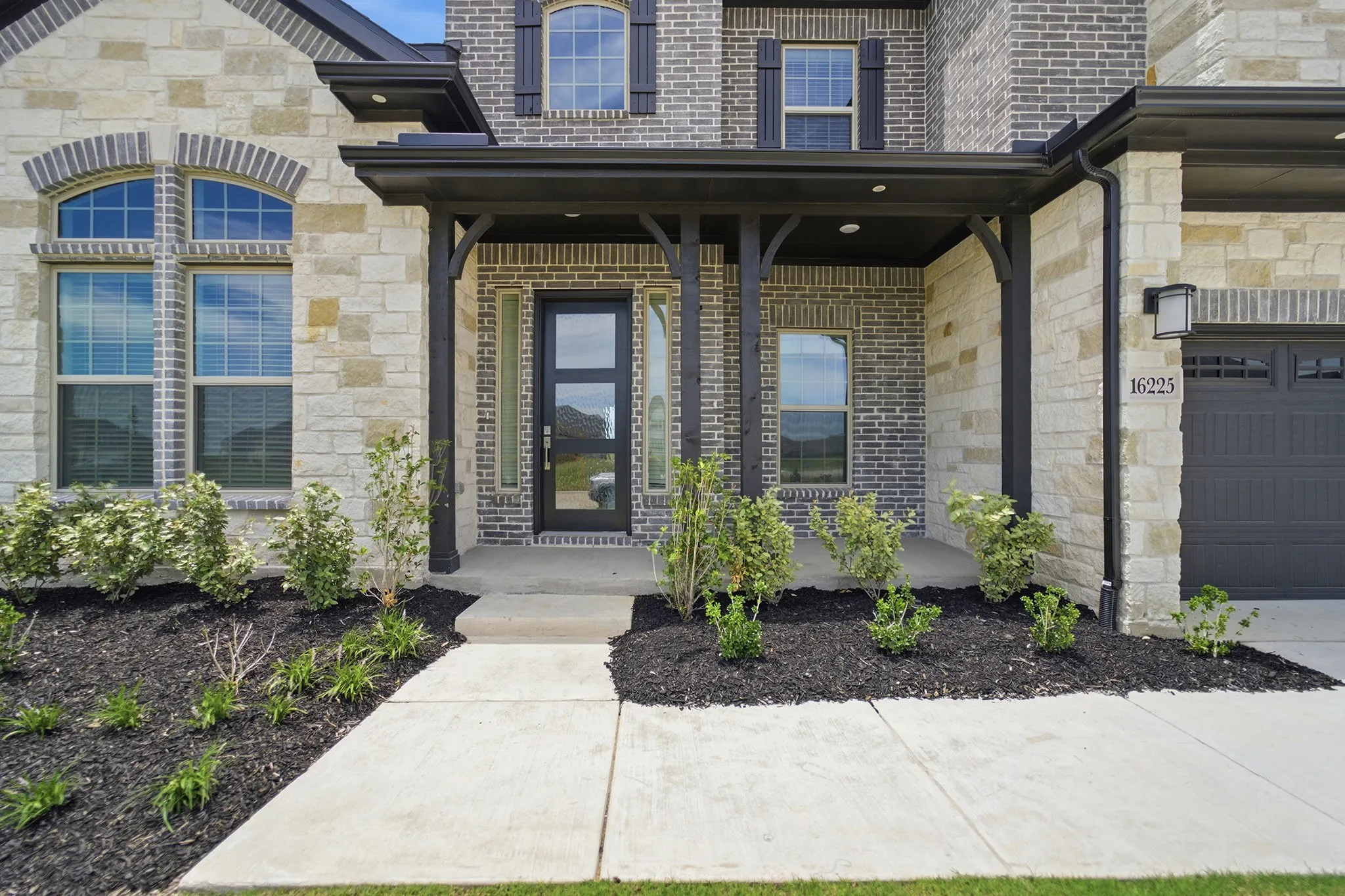 Two-story exterior featuring a grey brick facade with light-colored stone accents, a covered entry with dark wood-finish columns, a dark panel garage door, a modern glass front door, and manicured landscaping with dark mulch