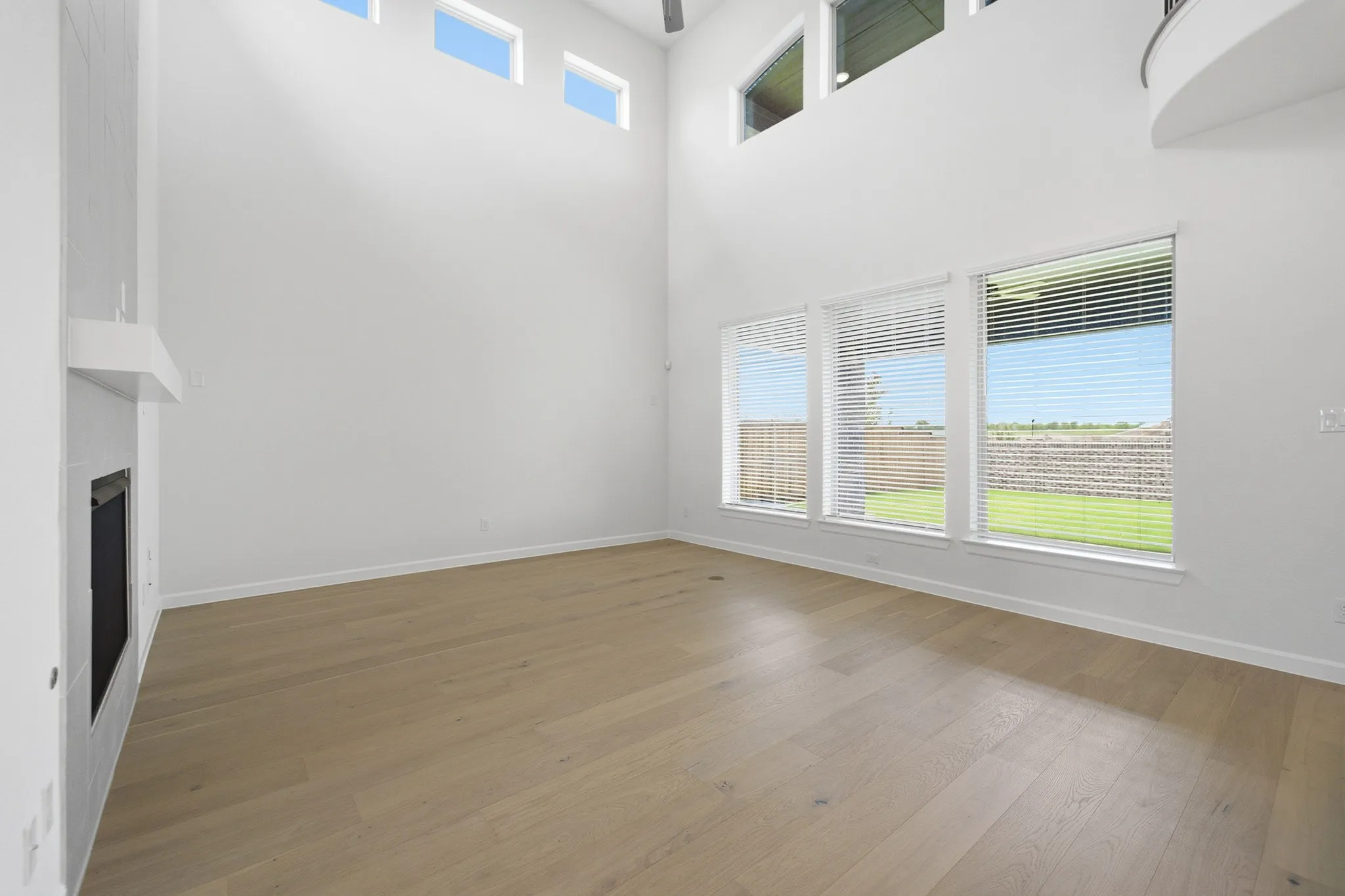 Spacious living area featuring high ceilings, wood-finish flooring, a contemporary fireplace, and multiple windows with blinds overlooking a green lawn