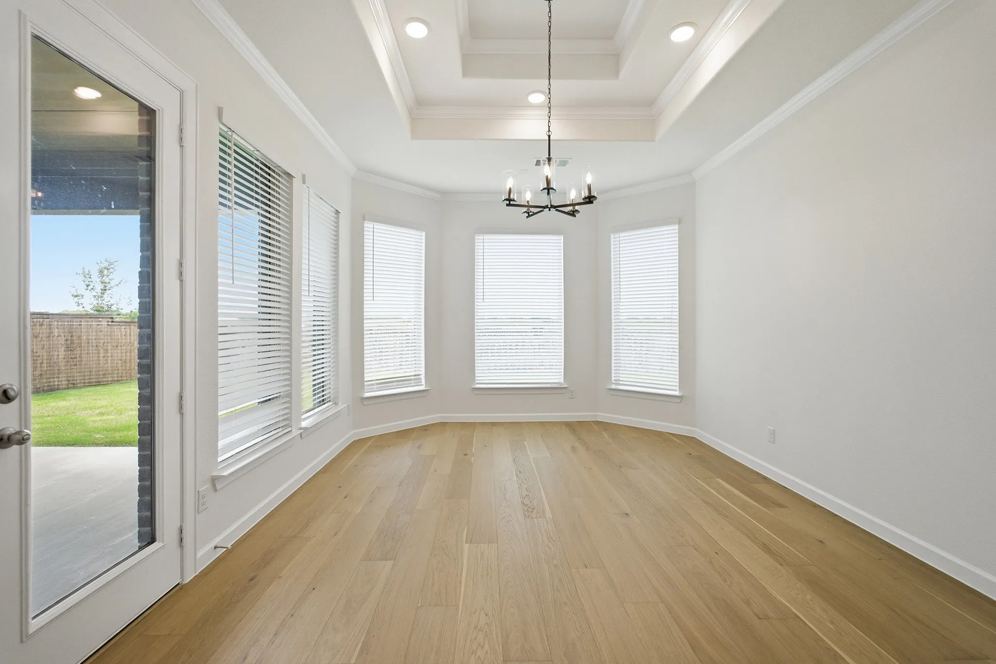 Spacious room featuring wood-finish flooring, a tray ceiling with recessed lighting, an iron chandelier, a bay window with blinds, and a glass-paneled exterior door