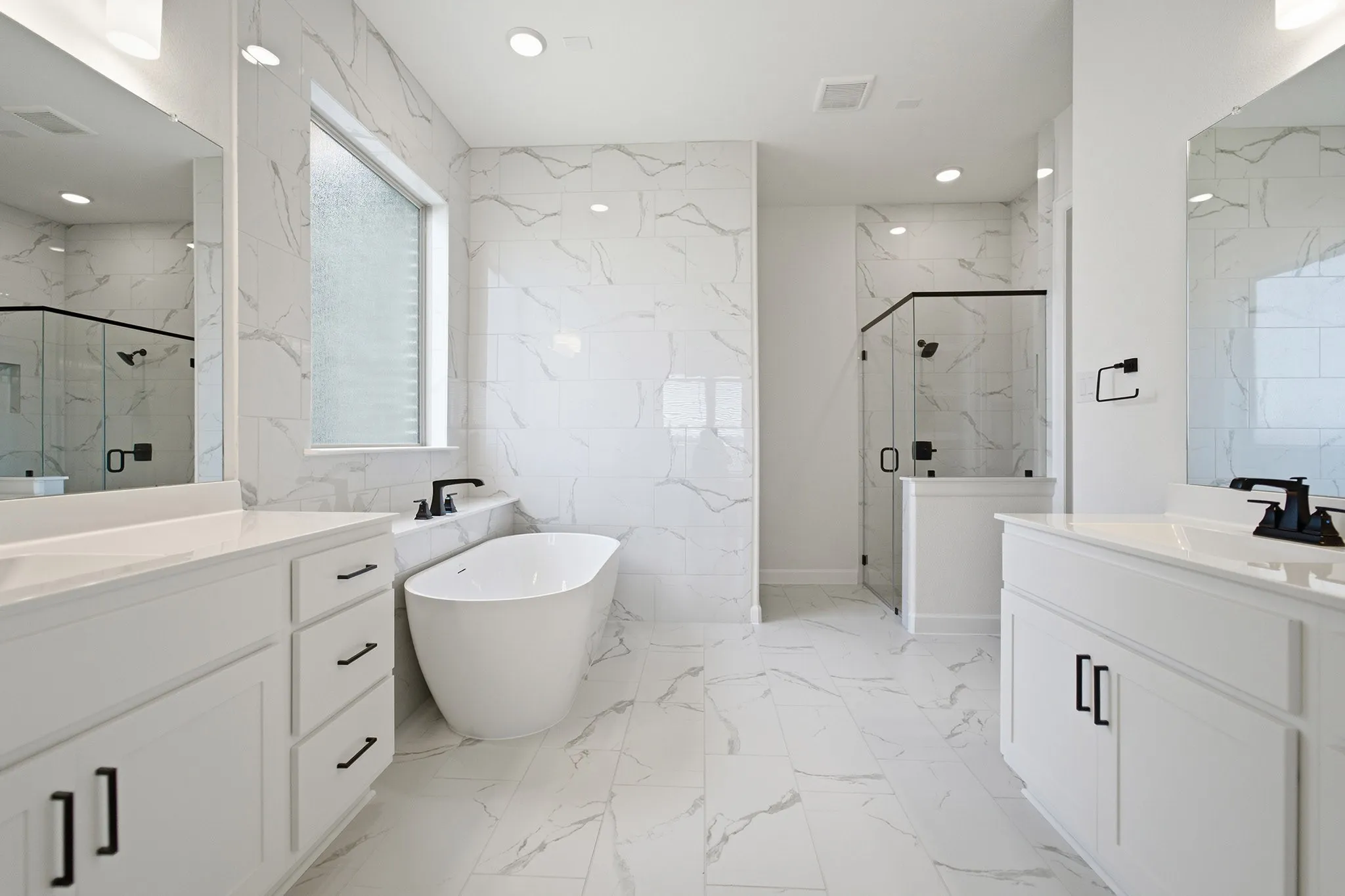 Spacious bath featuring a freestanding tub, dual vanities with white cabinetry, a glass-enclosed shower, and large format tile flooring
