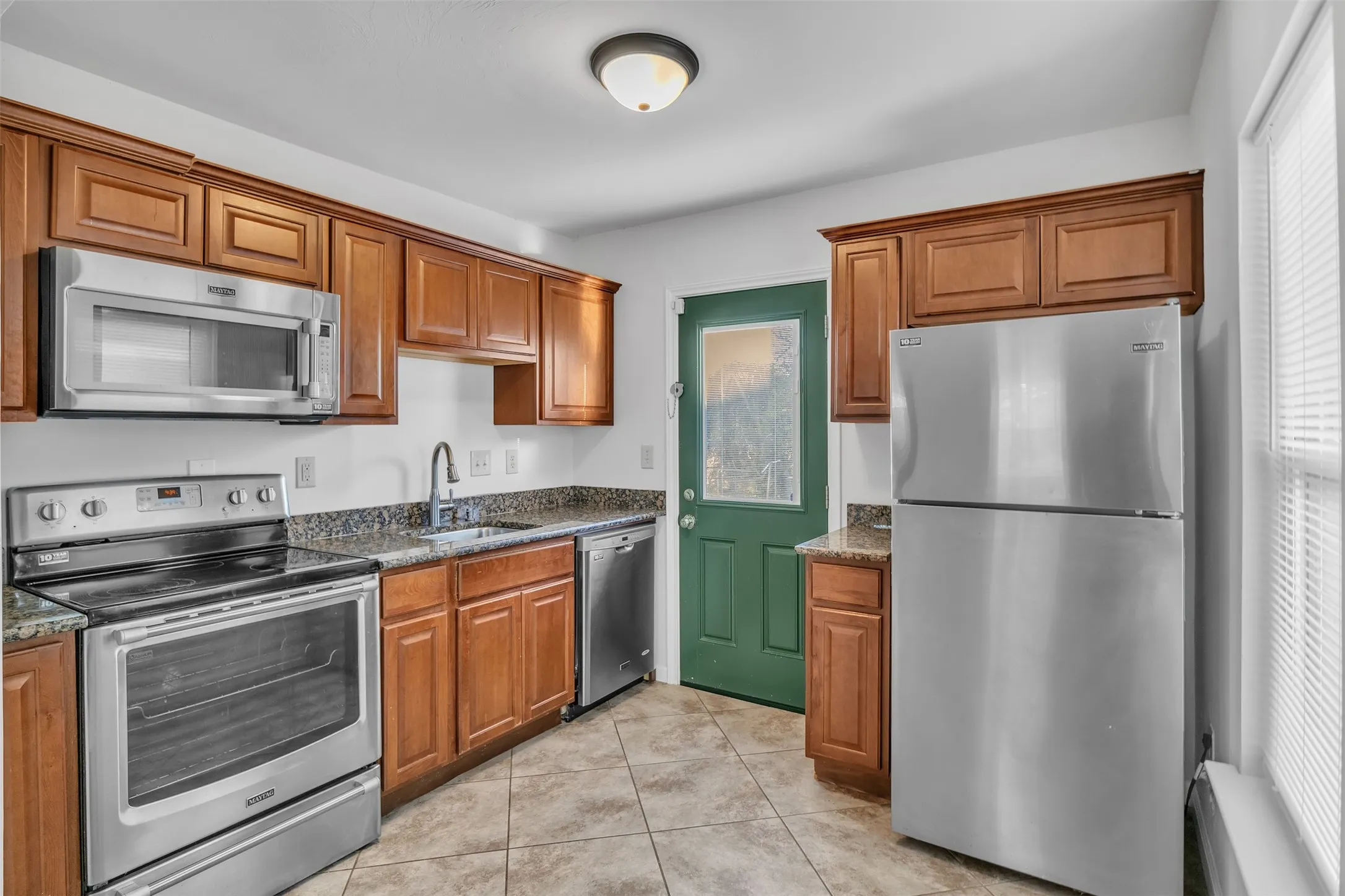 Kitchen with appliances with stainless steel finishes, brown cabinetry, light tile patterned floors, and dark stone countertops