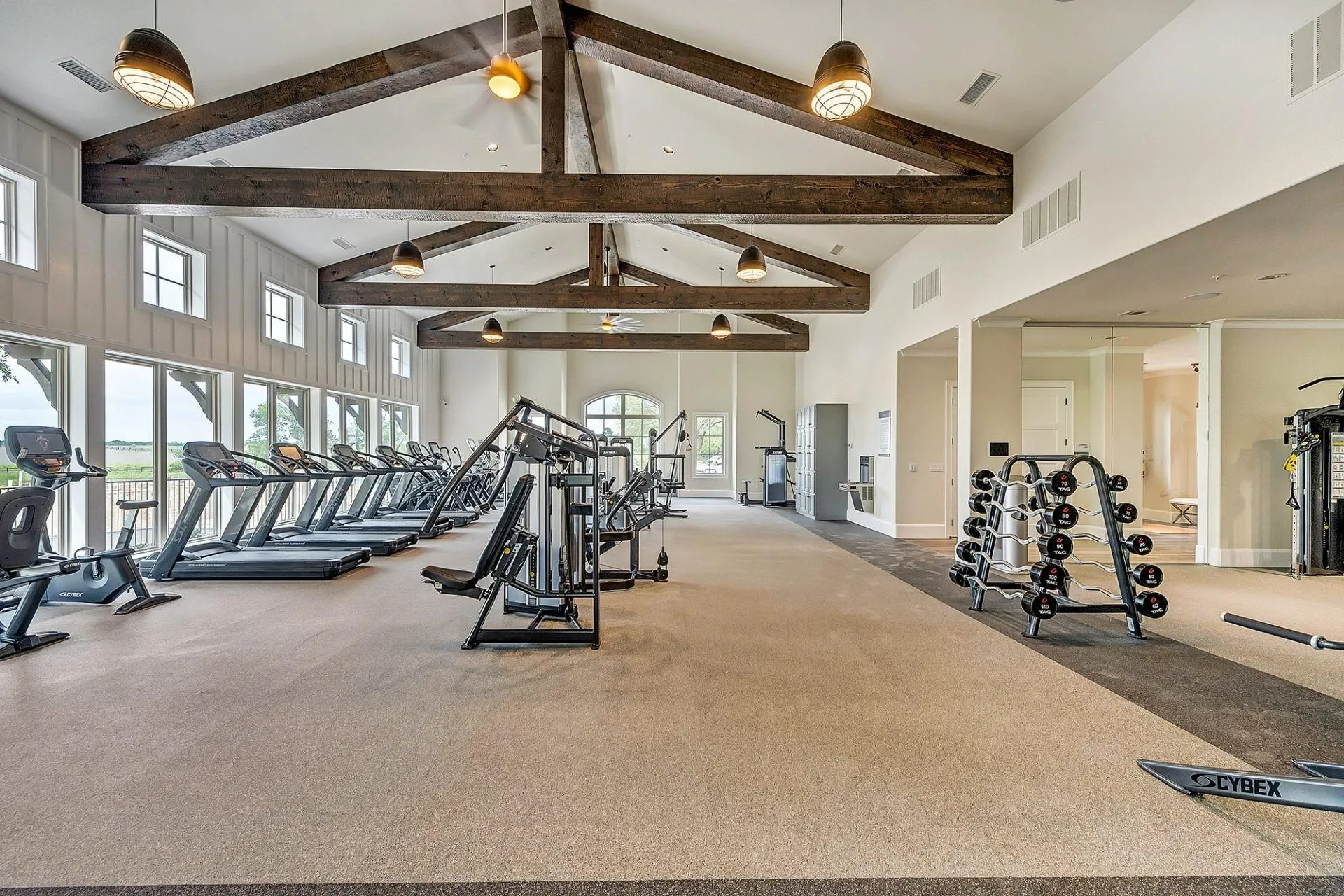 Spacious fitness center featuring vaulted ceilings with exposed wood beams, multiple large windows, a variety of cardio equipment, strength training machines, and a dedicated free weight area