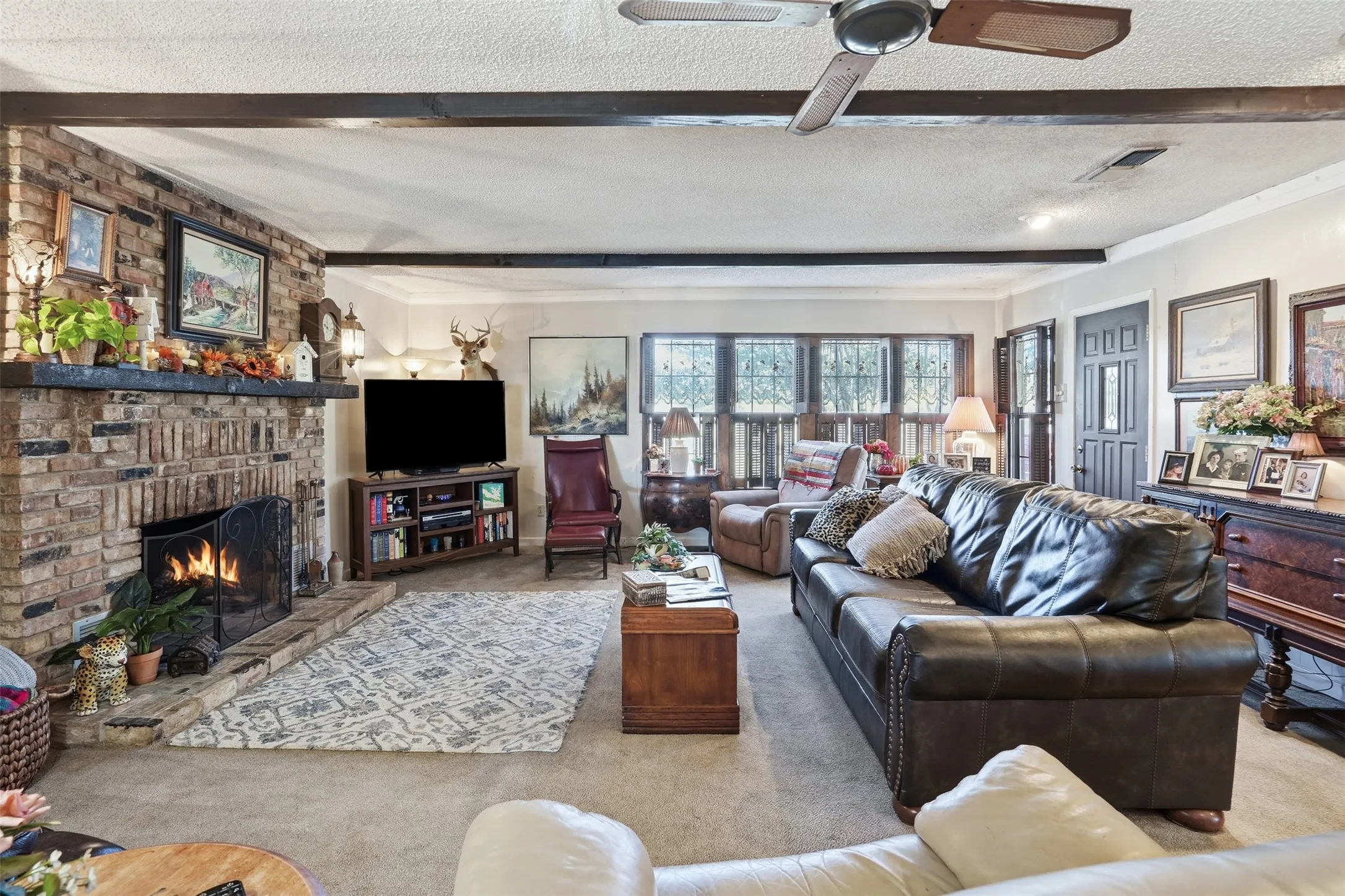 Living area with carpet floors, a textured ceiling, a brick fireplace, beam ceiling, and ceiling fan