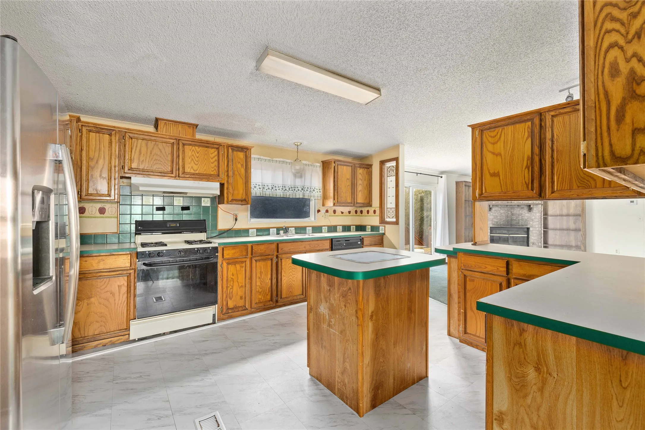 Kitchen with stainless steel refrigerator with ice dispenser, gas range oven, brown cabinetry, light countertops, and a textured ceiling