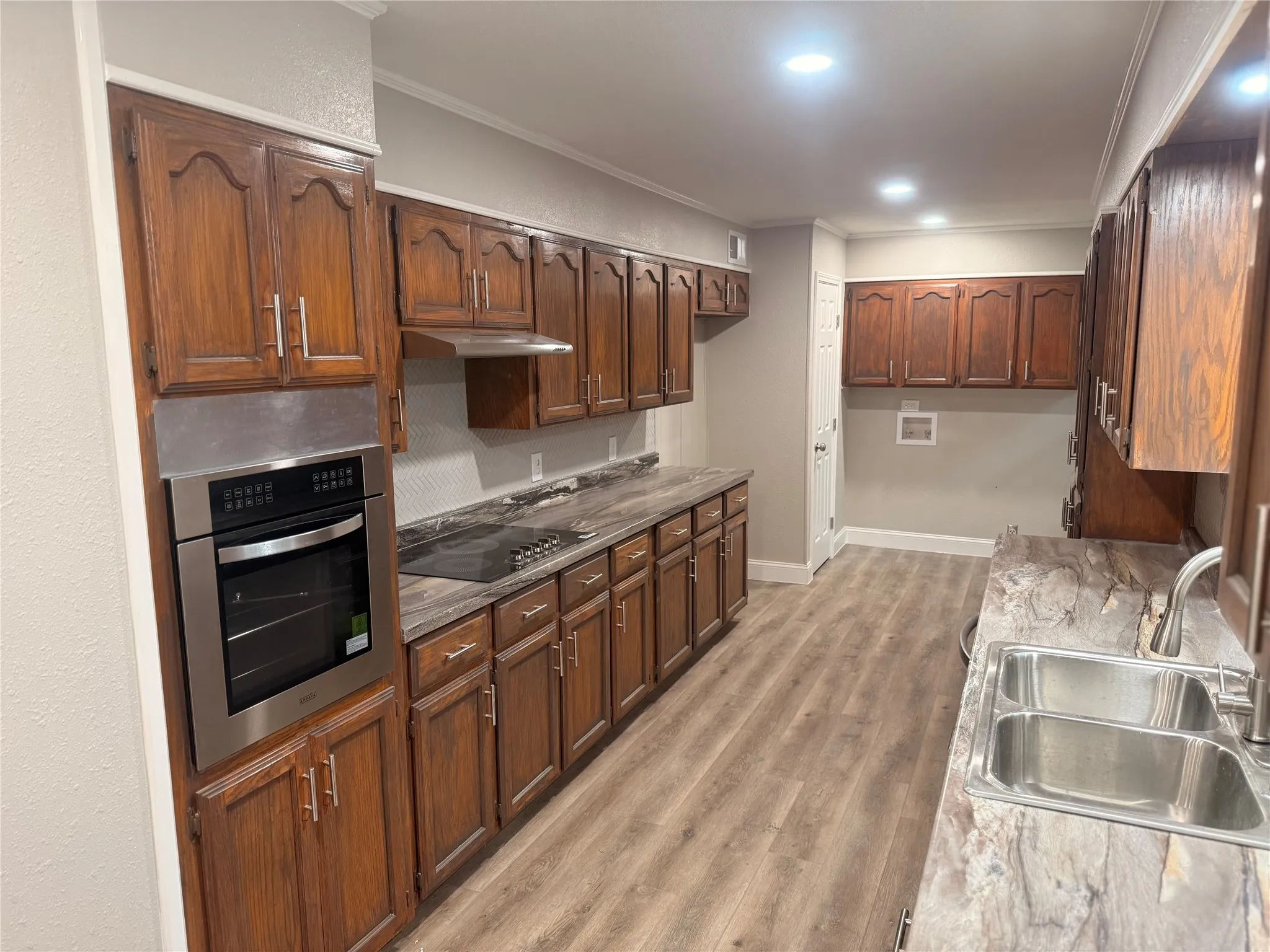 Kitchen featuring ornamental molding, stainless steel oven, light wood finished floors, black electric stovetop, and under cabinet range hood