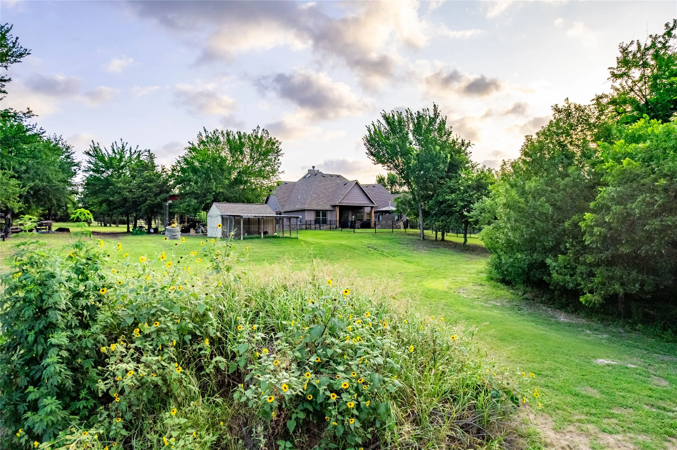 Expansive green lawn surrounds the property, featuring mature trees and a detached shed with an open-air enclosure