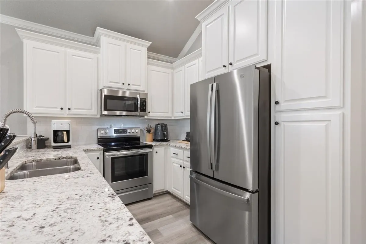 Kitchen with appliances with stainless steel finishes, light stone countertops, white cabinetry, vaulted ceiling, and backsplash