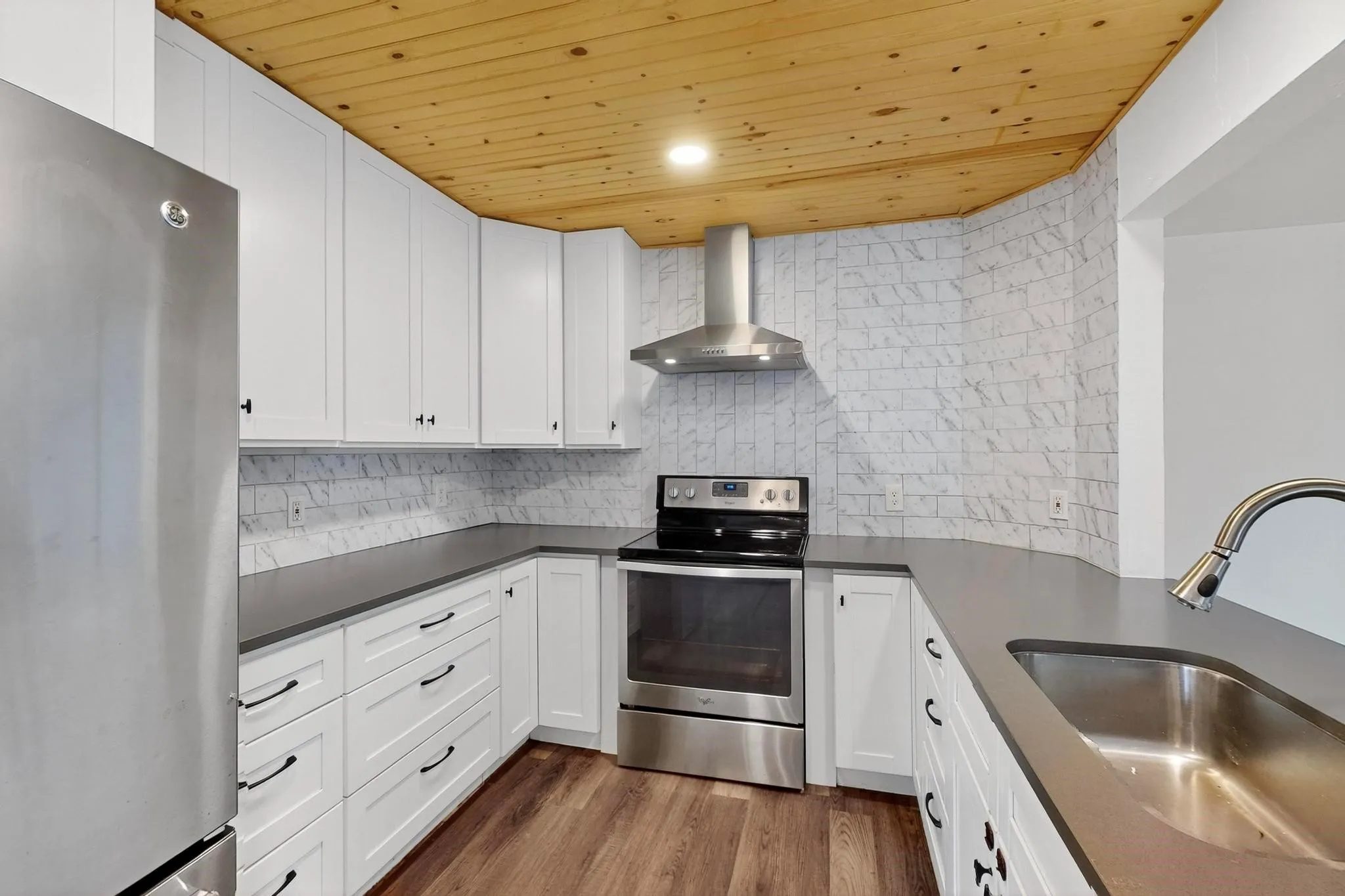 Kitchen with wooden ceiling, stainless steel appliances, wall chimney exhaust hood, white cabinets, and tasteful backsplash