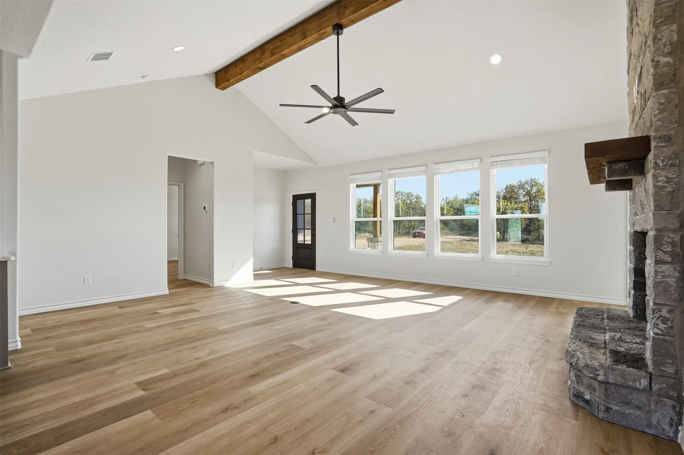 Unfurnished living room with a stone fireplace, light wood-style floors, high vaulted ceiling, a ceiling fan, and beam ceiling