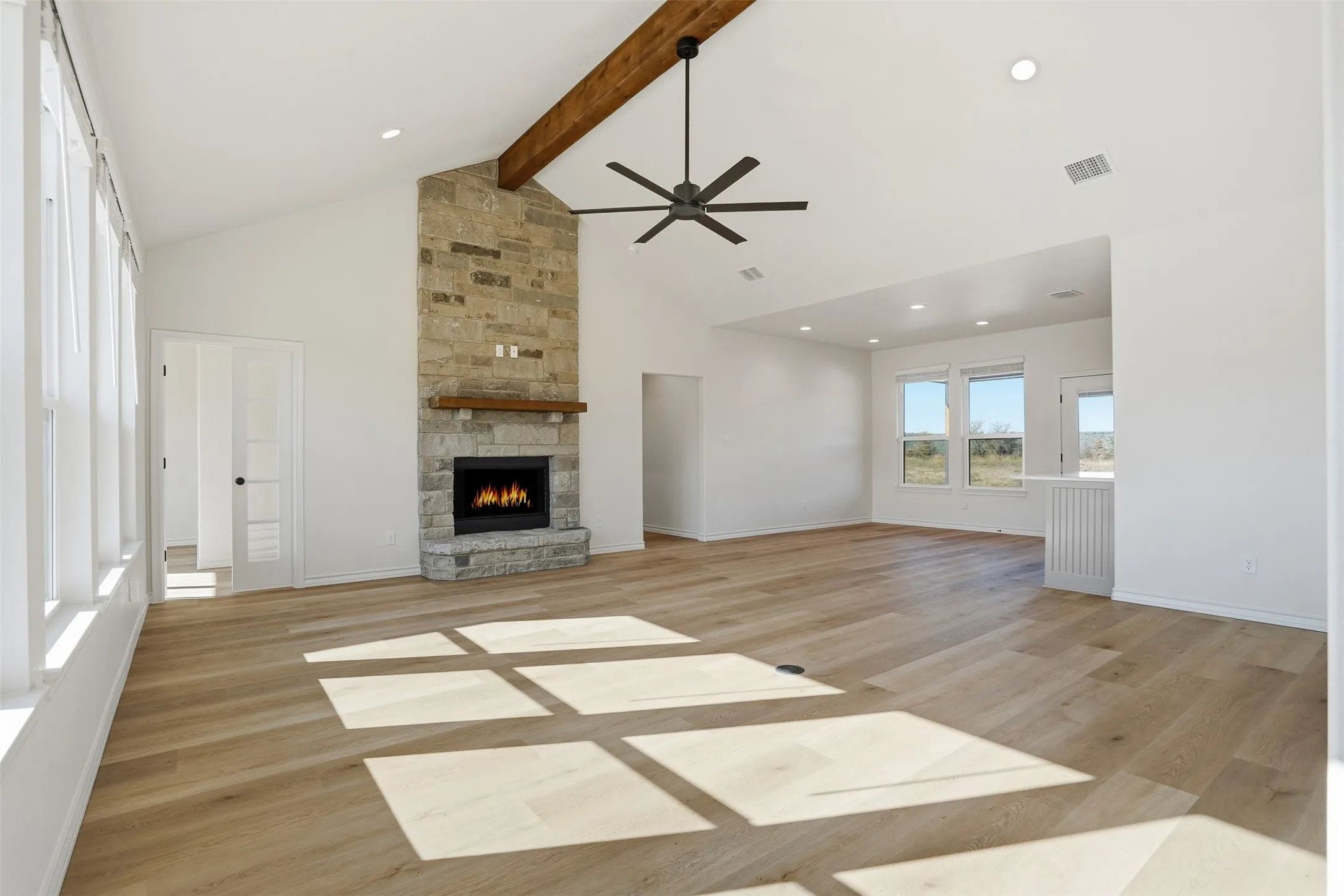 Unfurnished living room featuring a stone fireplace, a ceiling fan, light wood finished floors, high vaulted ceiling, and beam ceiling