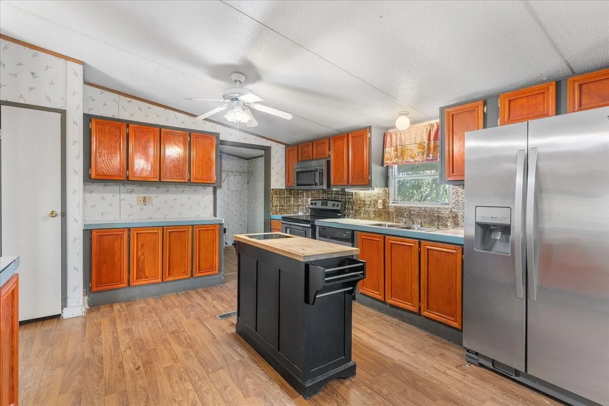 Kitchen with stainless steel appliances, vaulted ceiling, brown cabinetry, light wood-type flooring, and wallpapered walls