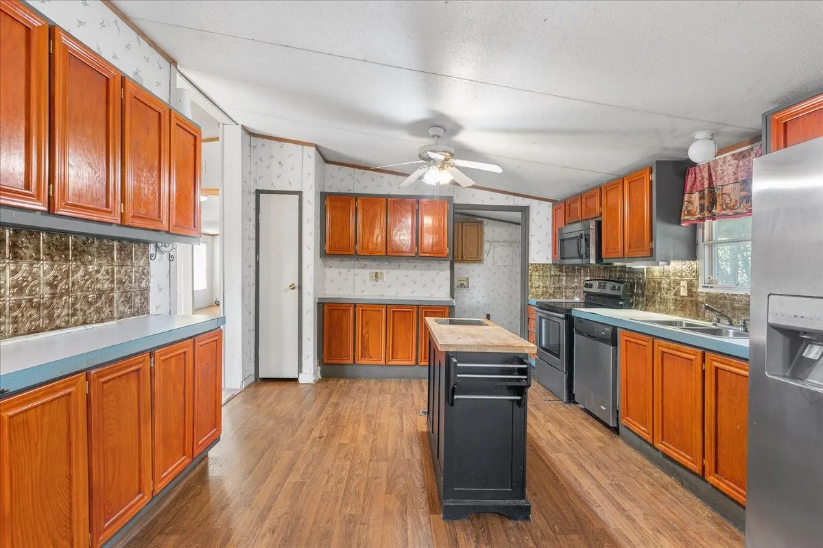 Kitchen featuring brown cabinetry, lofted ceiling, wallpapered walls, appliances with stainless steel finishes, and dark wood-type flooring