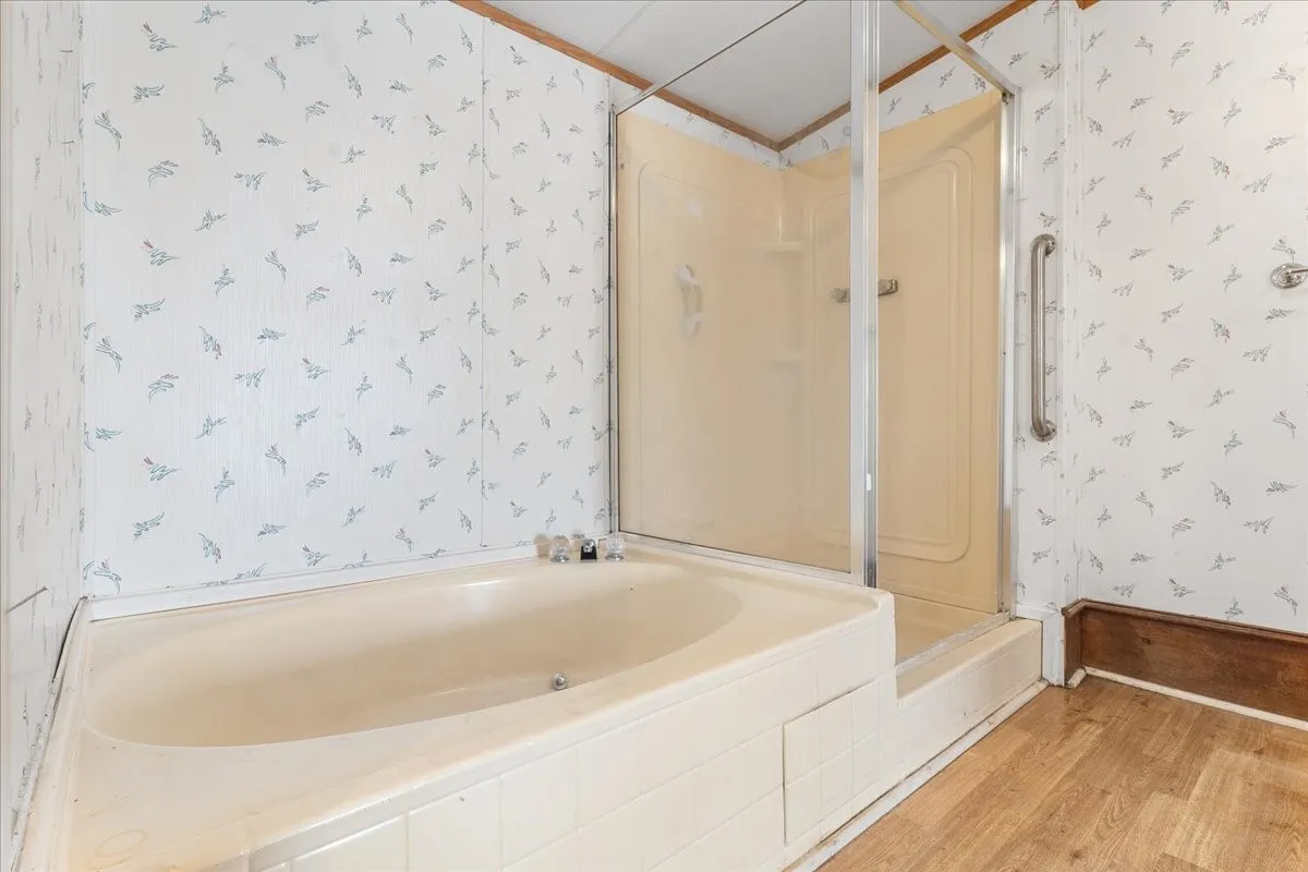 Bathroom with wallpapered walls, wood finished floors, a stall shower, and tiled bath